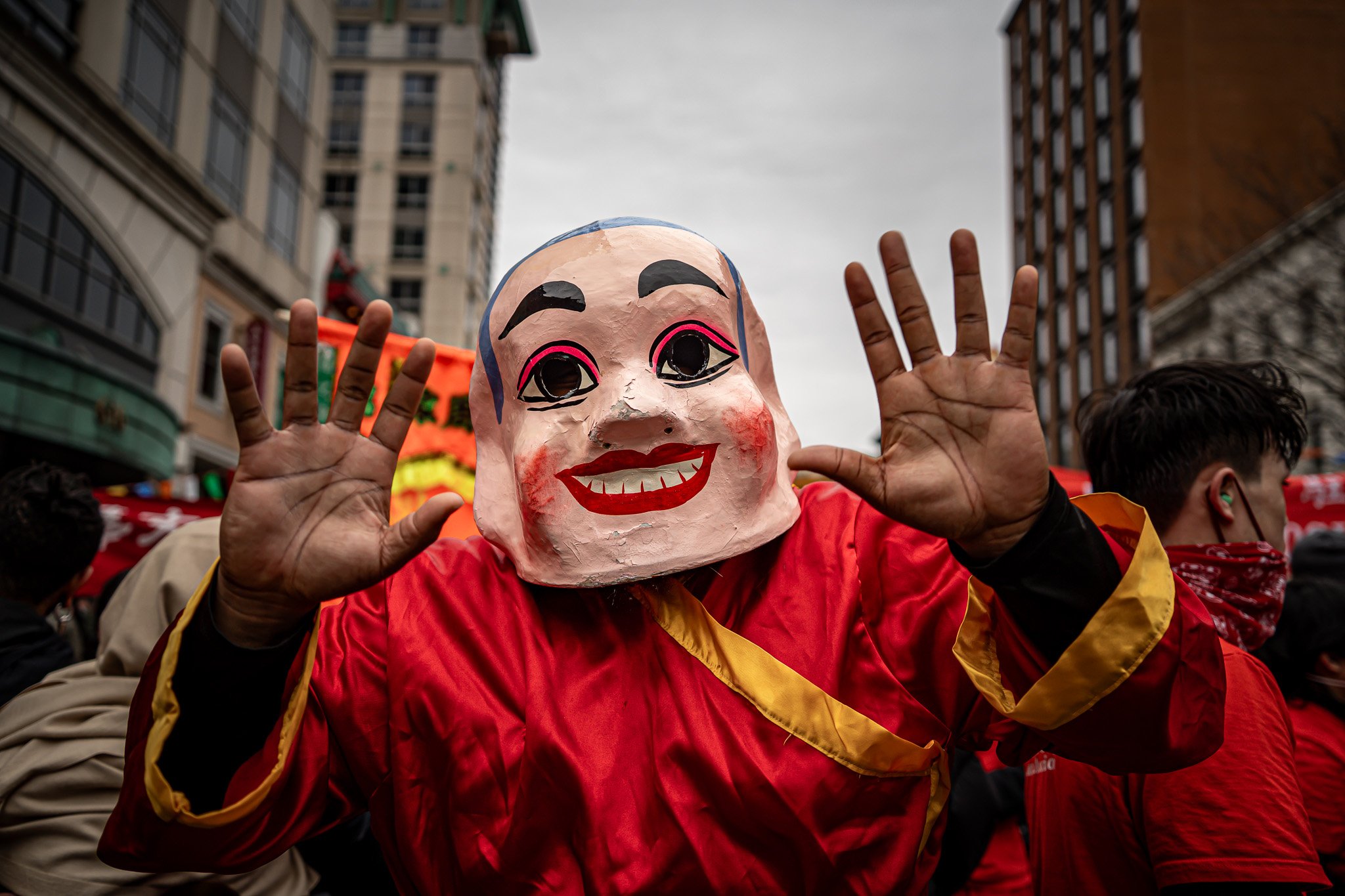 Person wearing a large, painted mask with exaggerated facial features and a big smile, holding hands up in a friendly gesture during a street event surrounded by buildings and other participants.