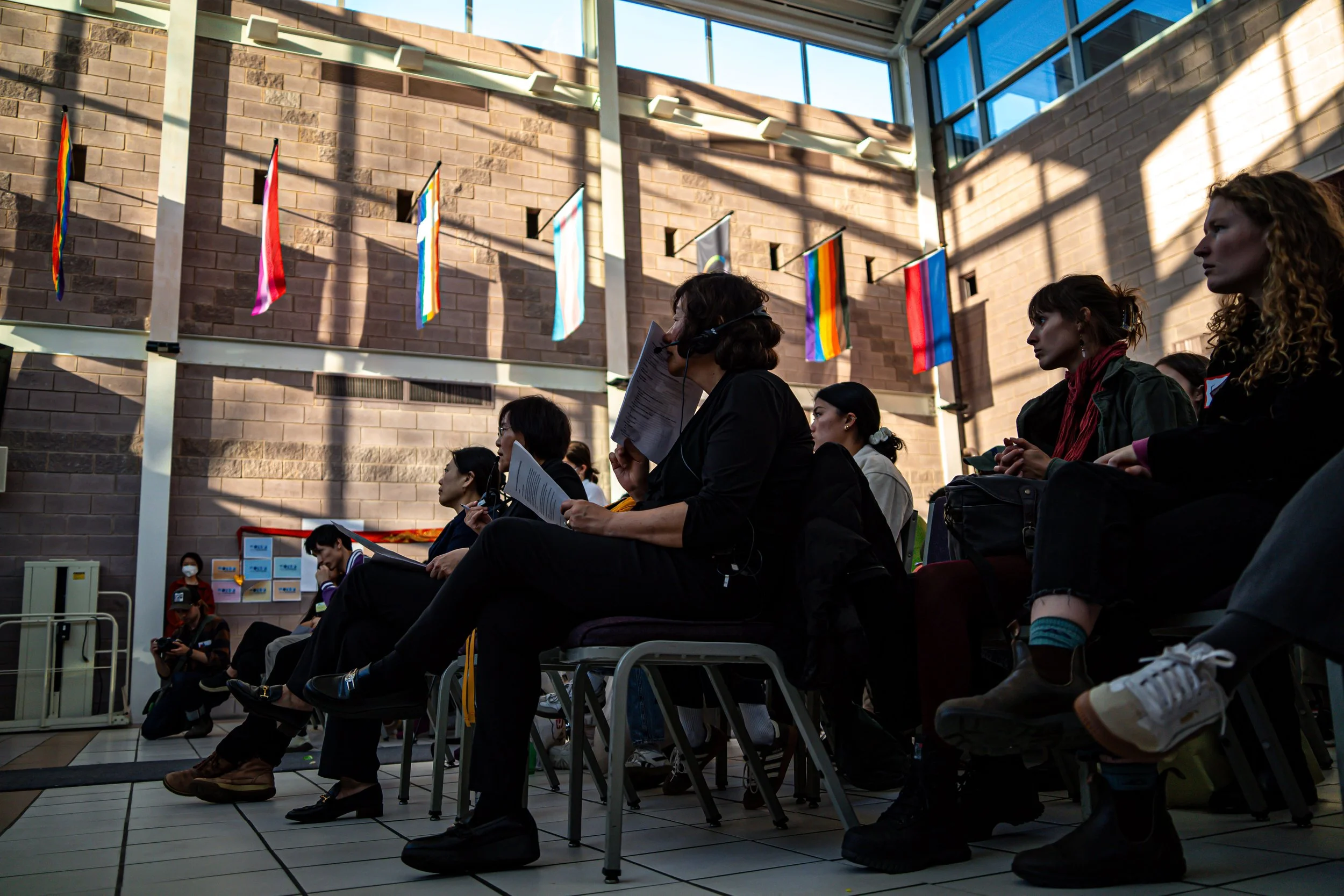 An audience at a conference or event, seated and listening. The room has rainbow pride flags hanging on the wall and large windows letting in natural light.