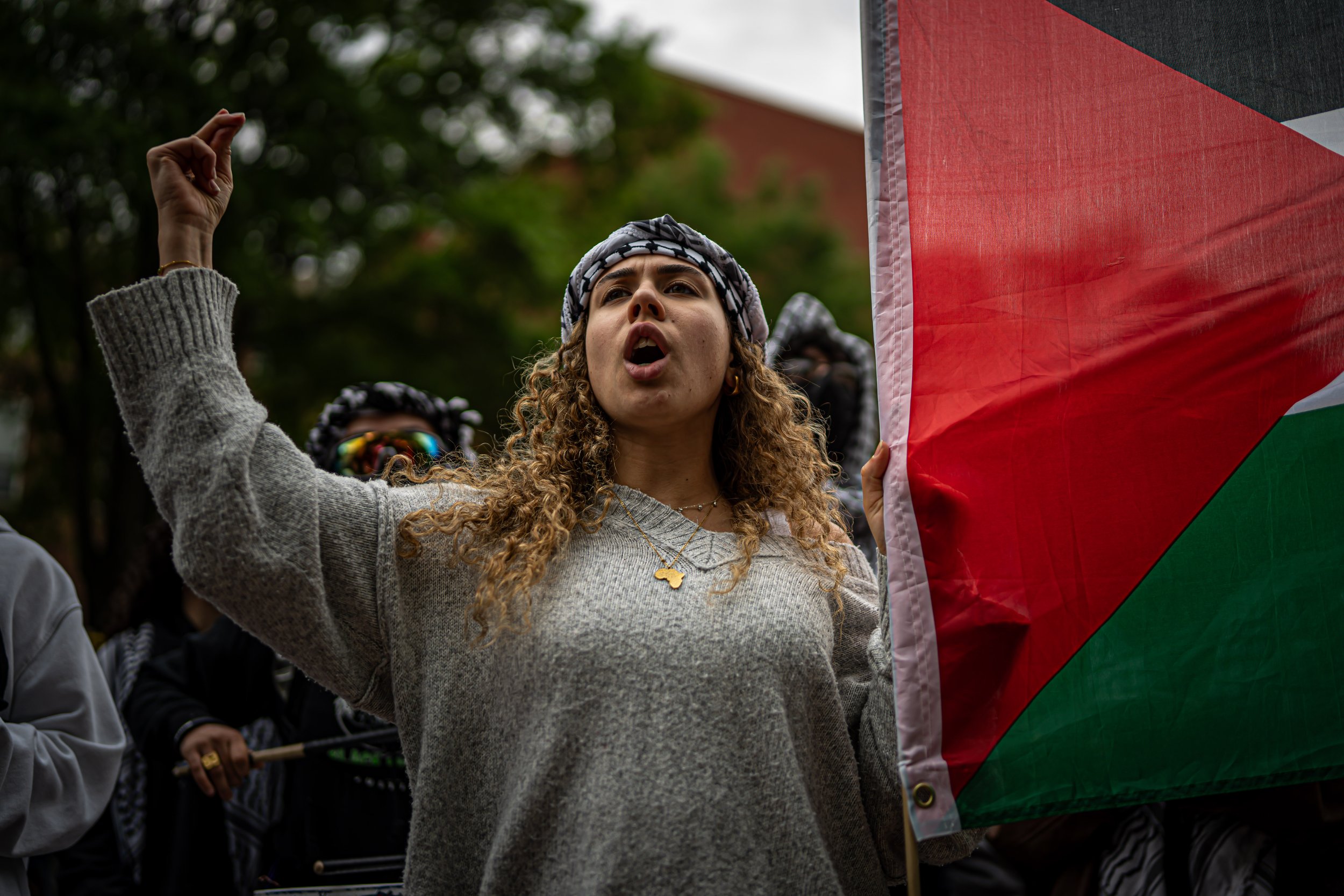 Woman protesting, holding a flag, wearing a headscarf, raised fist, shouting, surrounded by others.