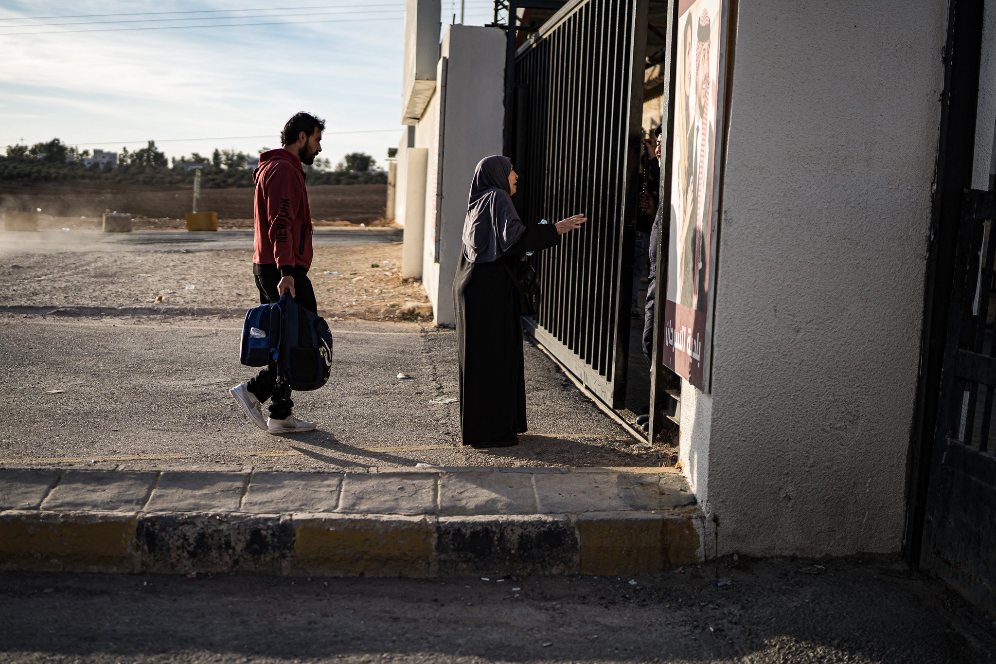 A woman in a dark hijab and dress is standing at a gate, reaching out to a person inside, while a man with a backpack stands behind her, waiting to enter, during daytime in a dry, open area.