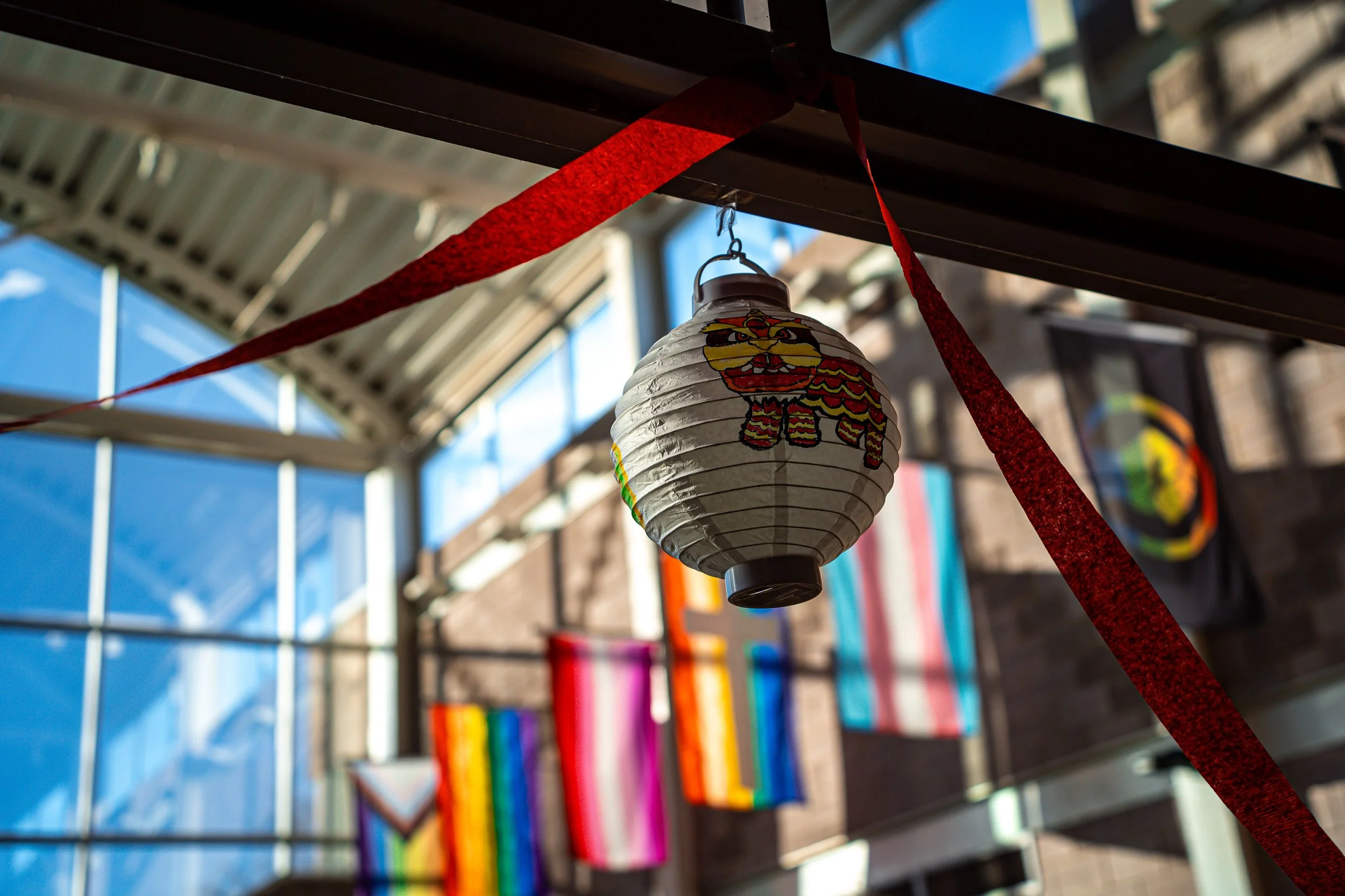 Paper lantern hanging from a red ribbon, with colorful rainbow flags in the background and sunlight streaming through windows.
