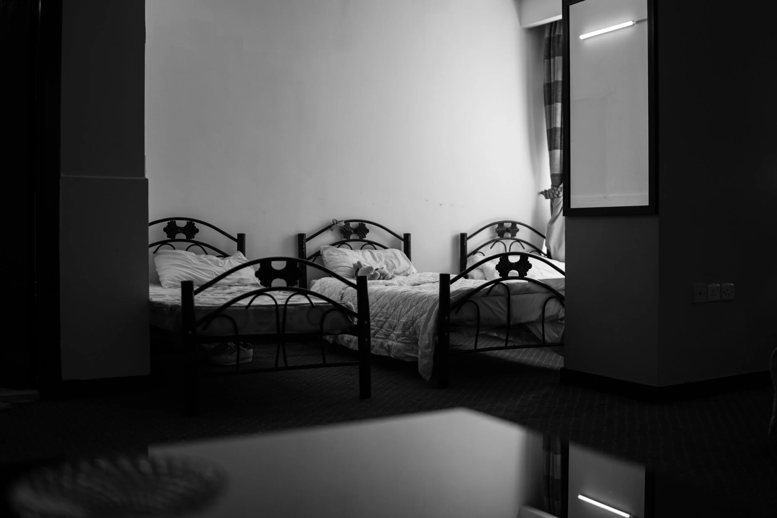 A black and white photo of a room with three beds, unmade with pillows and blankets, arranged in a row against the wall. There is a window with curtains to the right, allowing some light into the room.