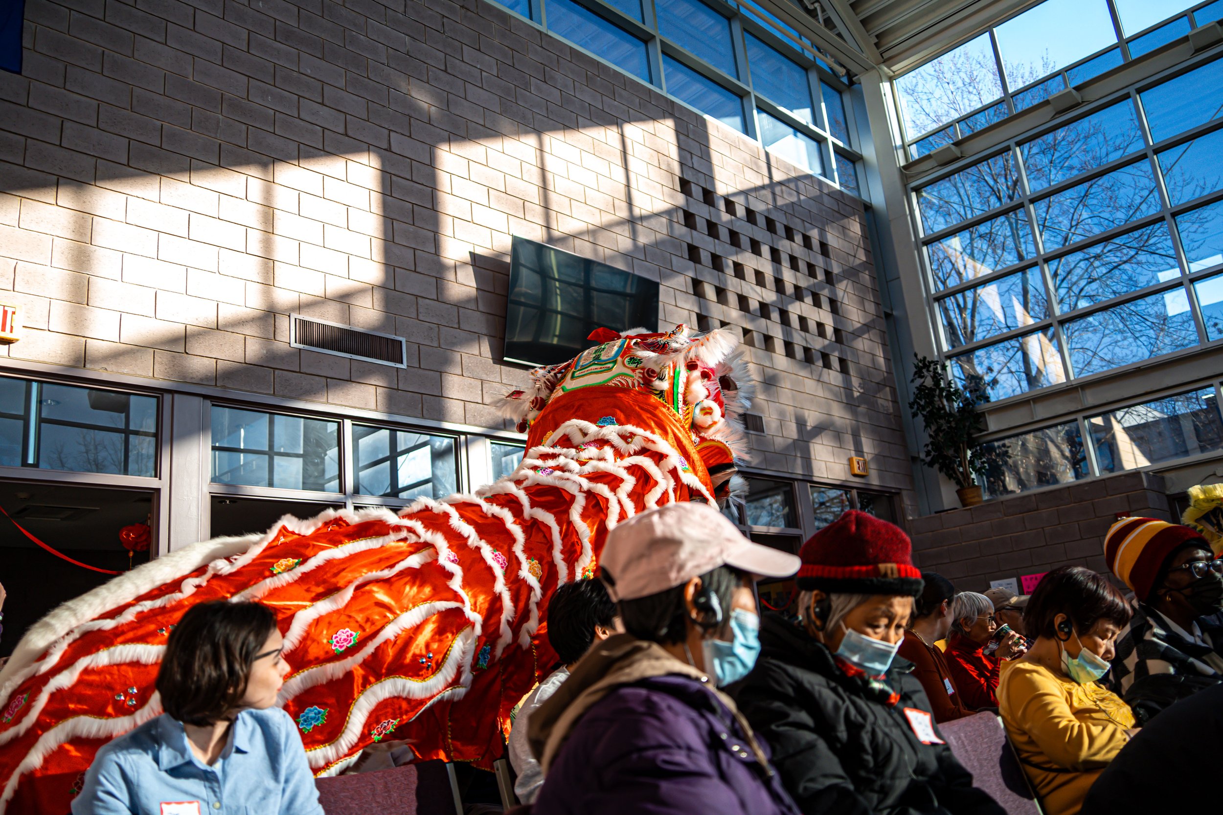 A traditional Chinese lion dance performance at an indoor event, with a bright orange lion costume decorated with white fur, colorful embroidery, and a character on the forehead, performed in front of seated individuals wearing face masks.