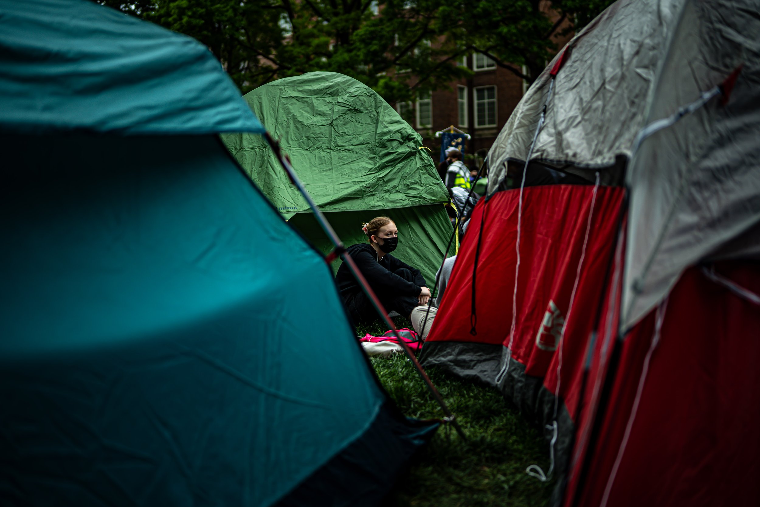 Person sitting on the grass surrounded by colorful camping tents, wearing a black mask and jacket.