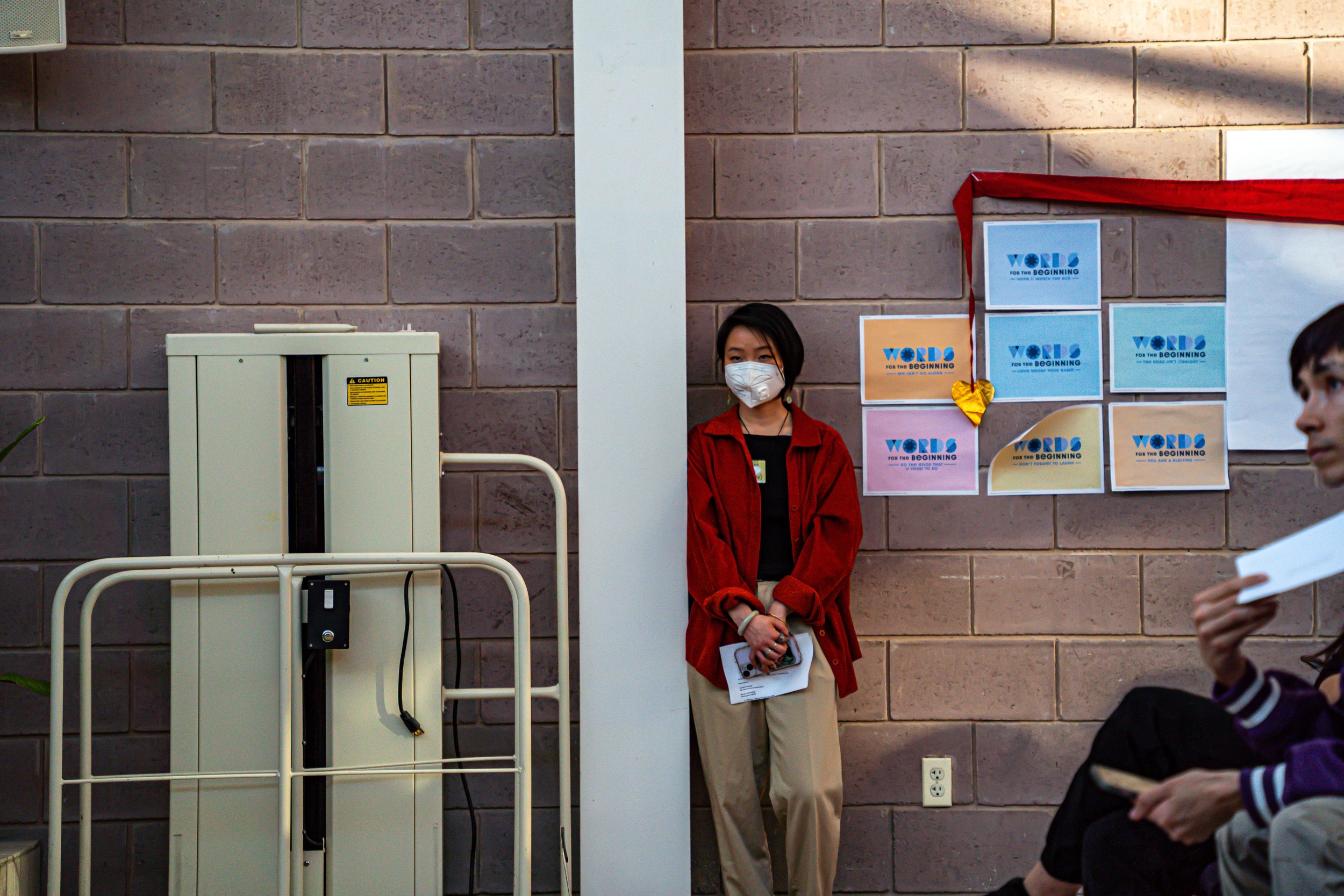 A woman with black hair wearing a face mask, a red jacket, and beige pants, standing against a brick wall next to a white pillar and a beige electrical box. She is holding papers and appears to be waiting. To her right, there are colorful posters on