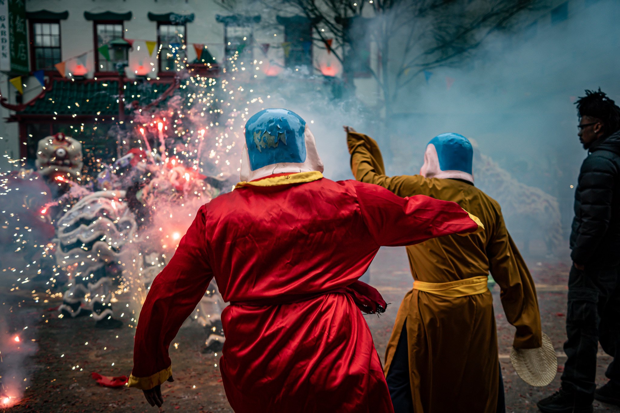 People in costumes with blue masks participating in a celebration with fireworks and decorative elements, possibly a festival.