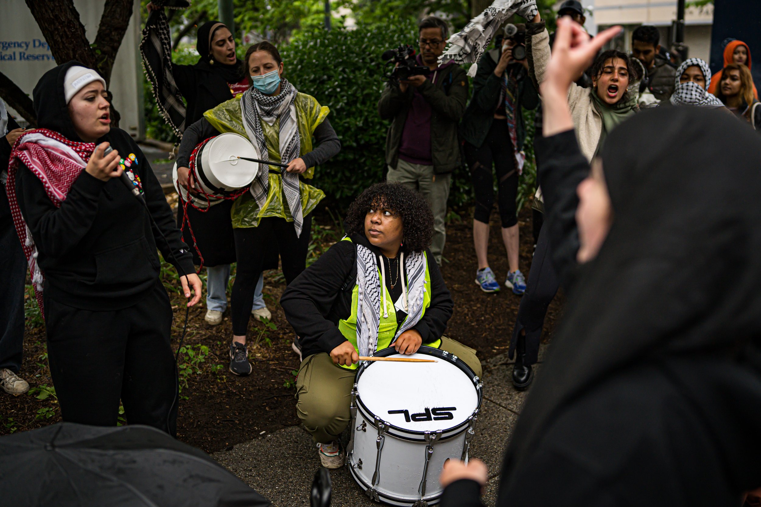 A group of people gathered outdoors, some singing and playing drums, with others taking photos. A woman in the center is sitting on the ground holding a drum, and a woman on the left is singing with her eyes closed. The atmosphere appears lively and