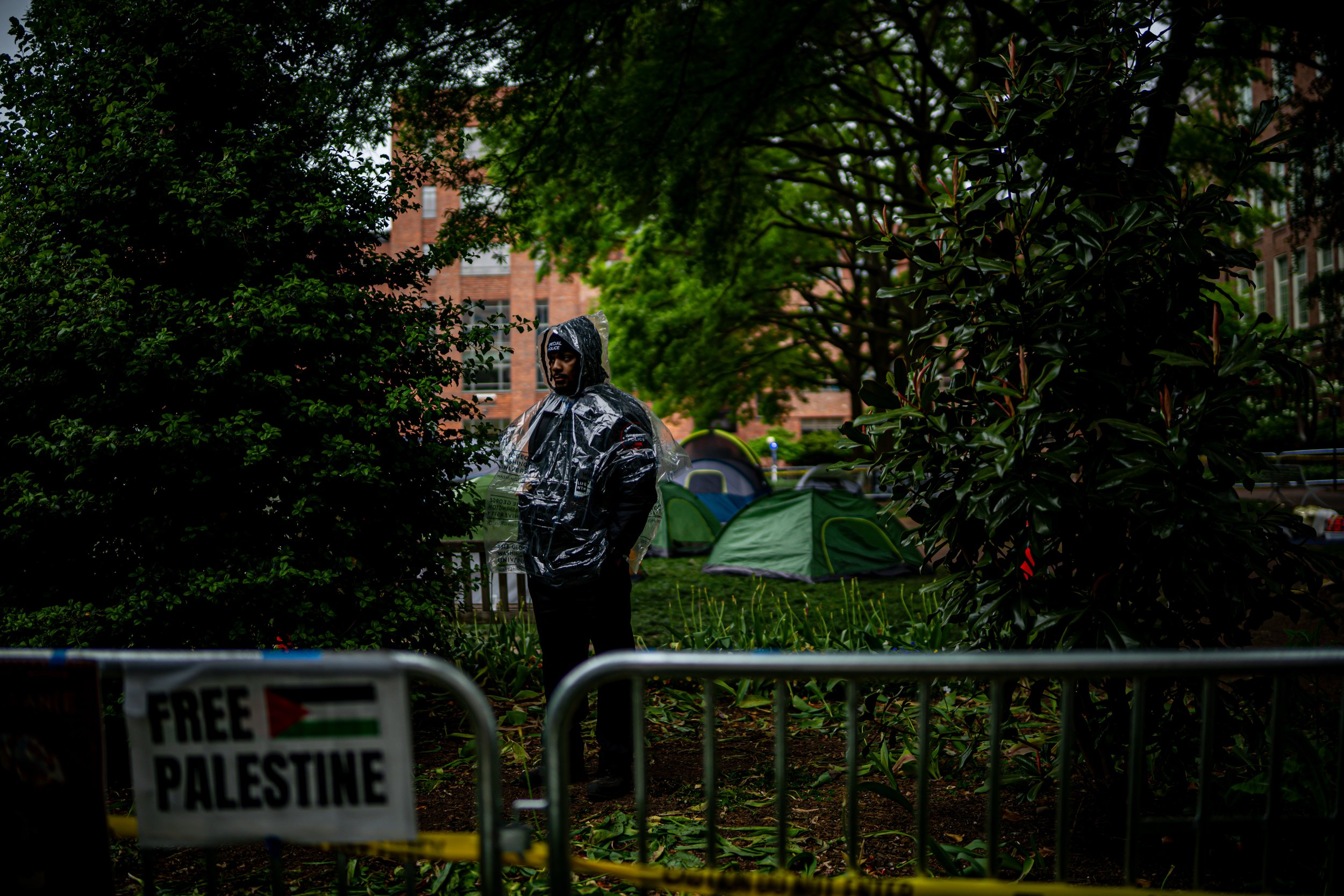 A man in rain gear standing in a park with tents in the background, behind a barrier with a 'Free Palestine' sign.