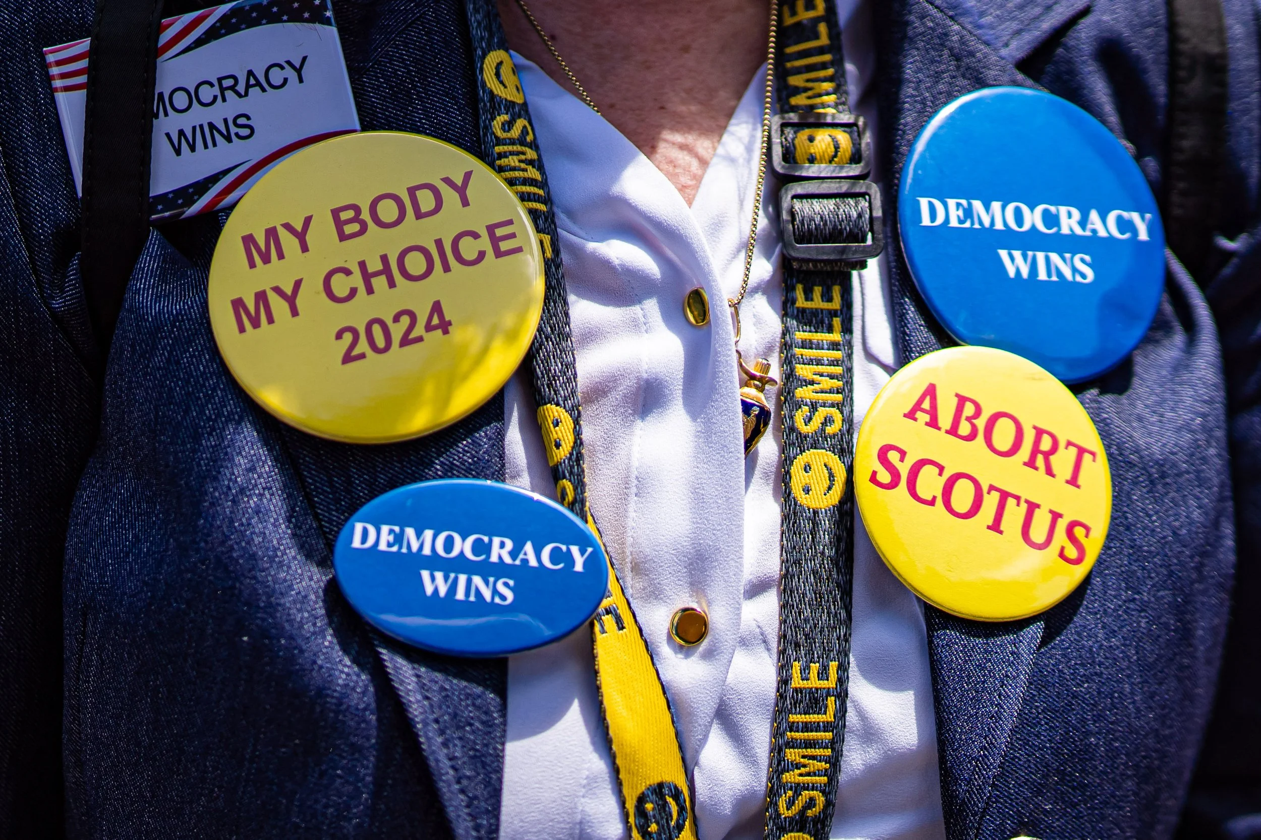 Collection of political buttons on a jacket, including yellow buttons that say 'My Body My Choice 2024' and 'Abort Scotus,' and blue buttons that say 'Democracy Wins'.