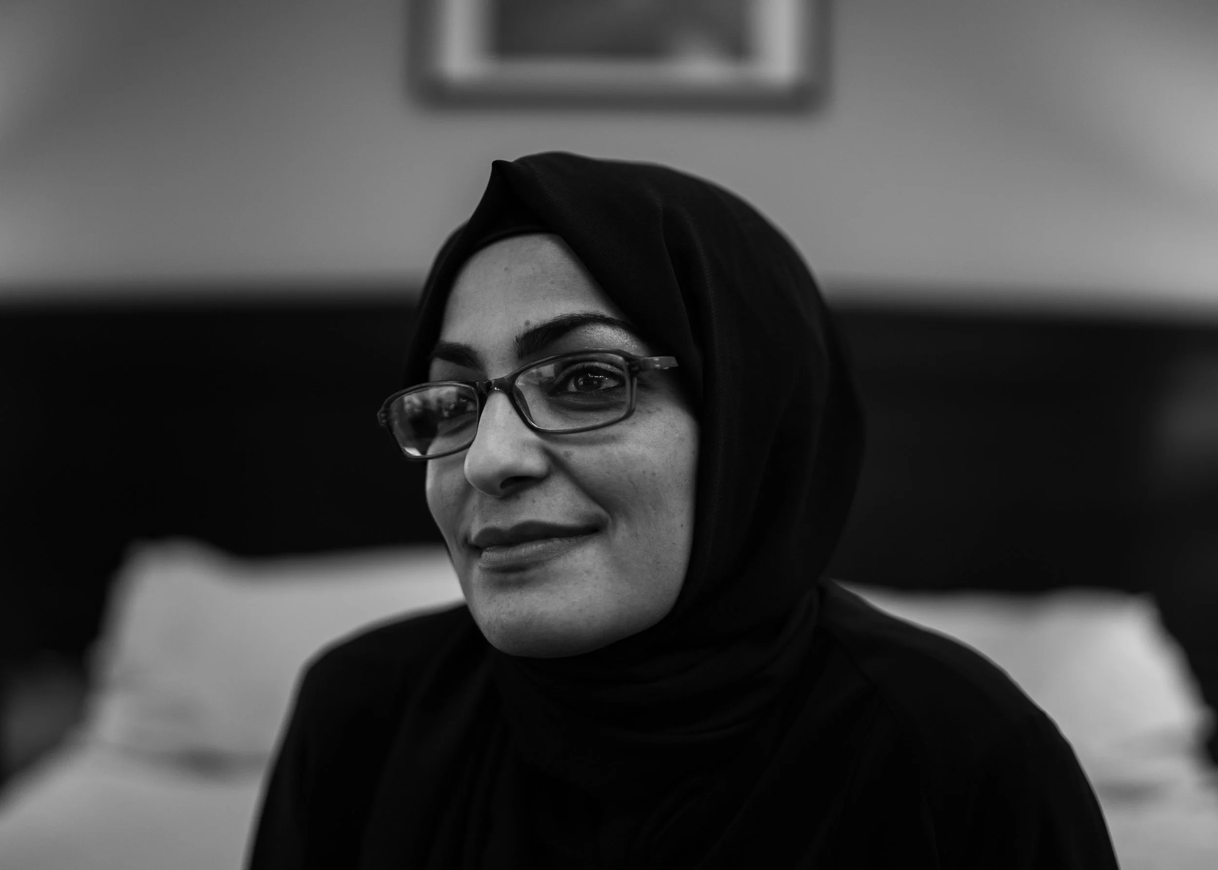 Black and white photo of a woman wearing glasses and a headscarf, smiling softly while looking slightly to the side.