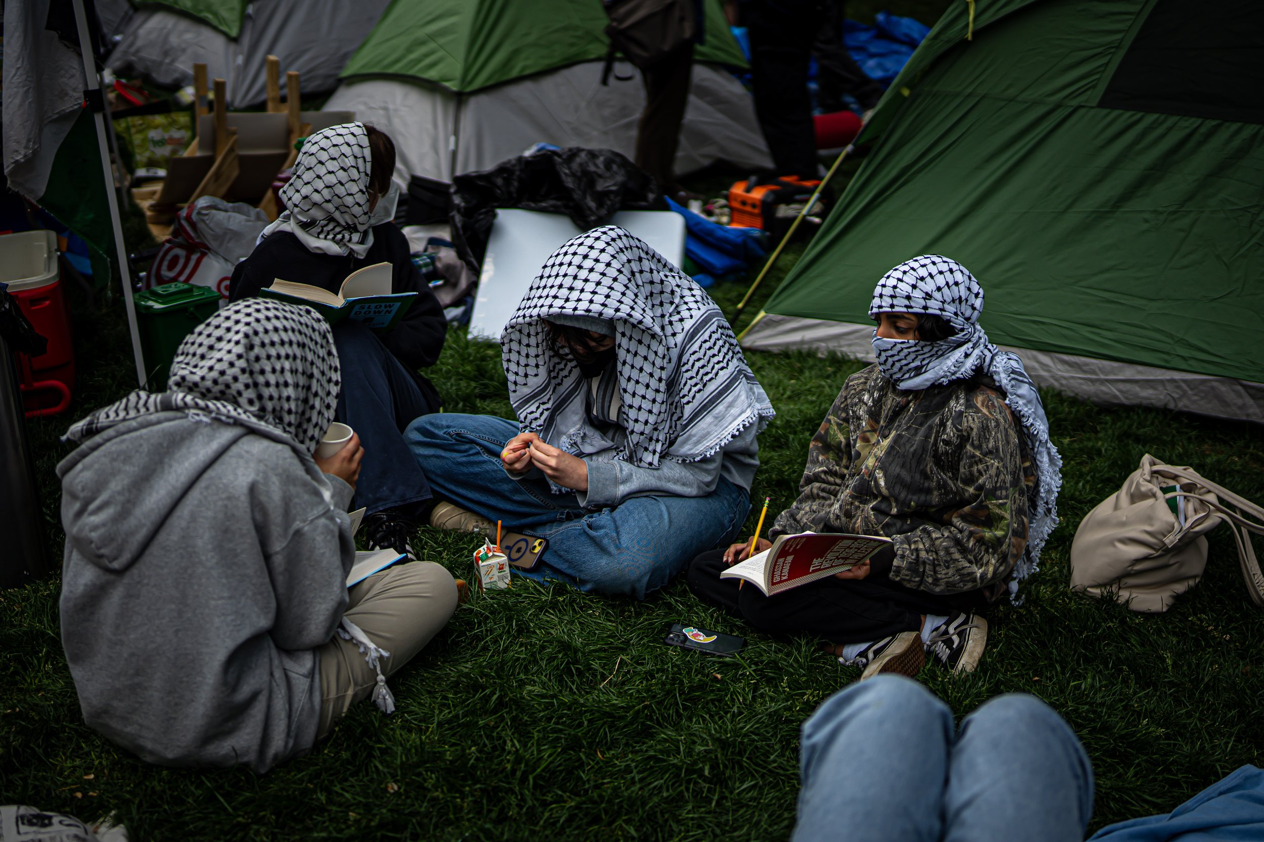 Four individuals wearing Palestinian keffiyeh scarves sit on grass in front of tents, reading, writing, and talking at a camping site.