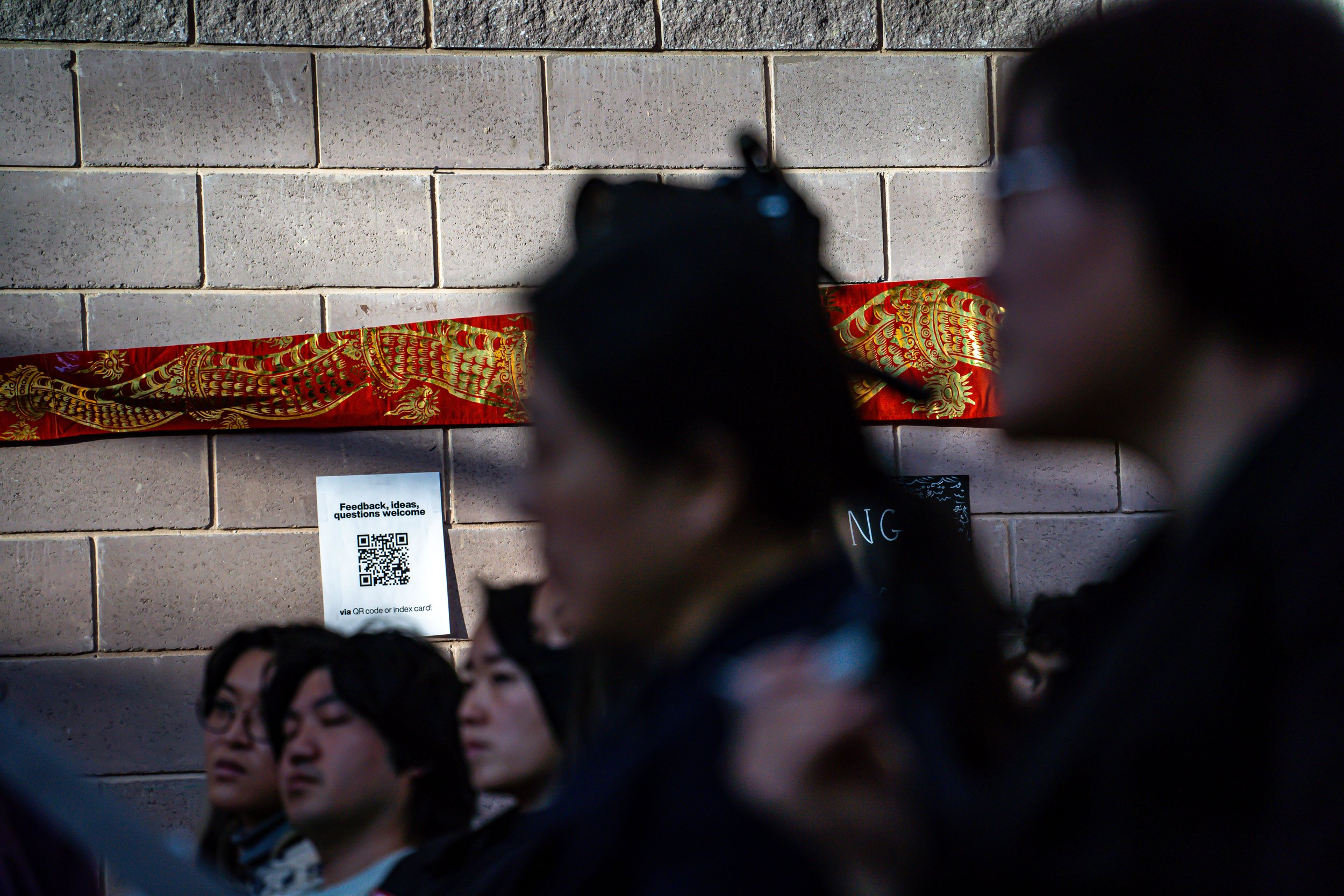 A group of people sitting in front of a brick wall with a red and gold decorative cloth and a sign with a QR code.