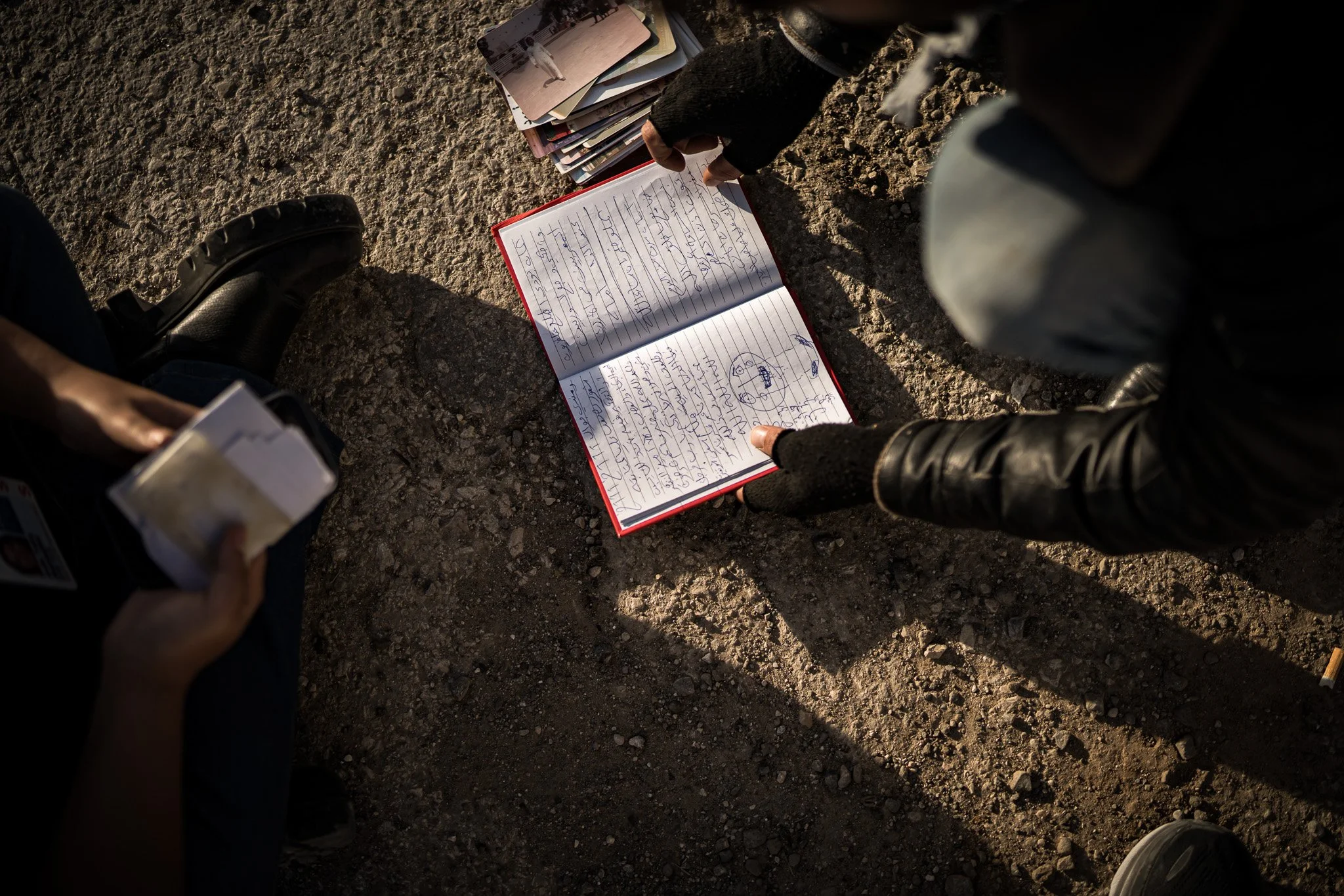 Two people are sitting on rocky ground, looking at an open red notebook with handwritten notes and drawings. One person is holding a small stack of papers or photos, and there are additional notebooks or folders nearby. The scene is lit by natural su