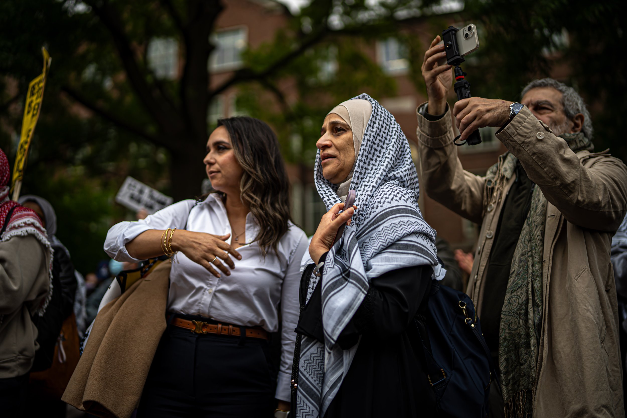 A diverse group of people participating in a protest or demonstration outdoors, with some holding signs and one person recording with a camera. The scene takes place in a park-like setting with trees in the background.