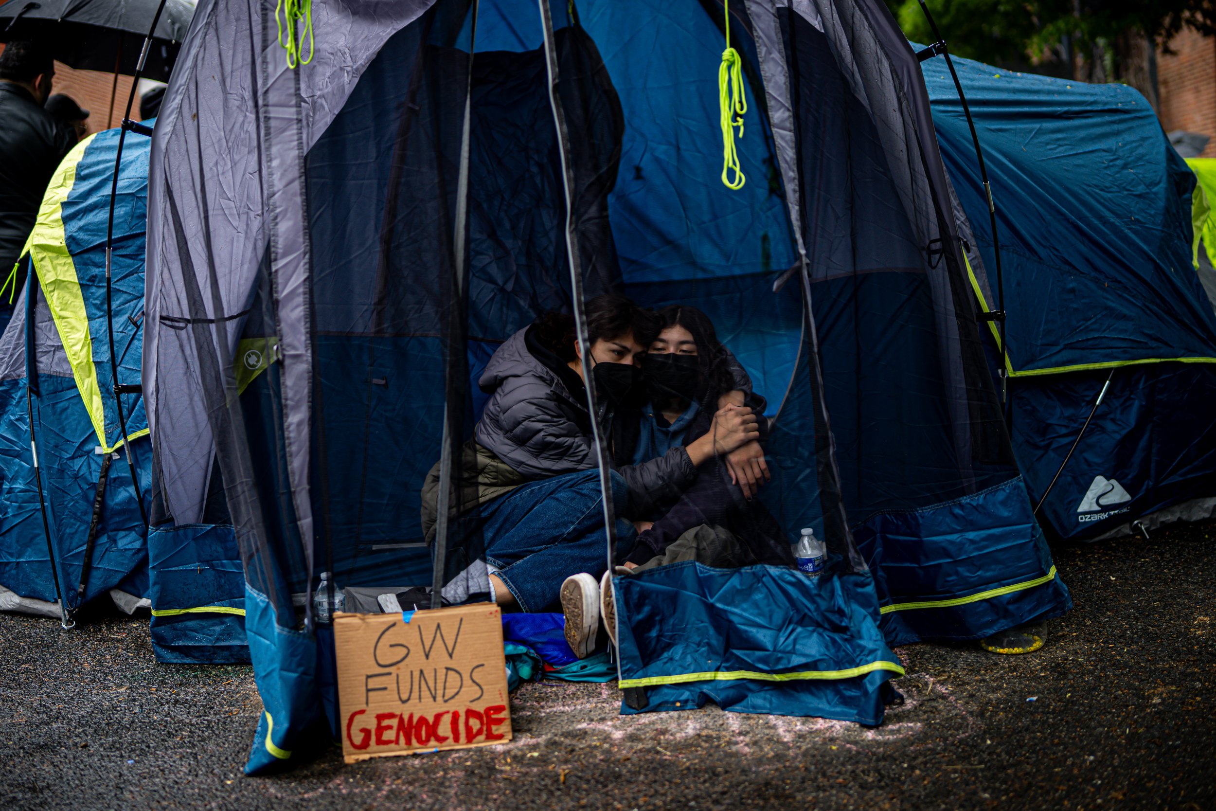 Two people sitting inside a tent at a protest, wearing masks, with a sign outside that reads 'GW Funds Genocide'.