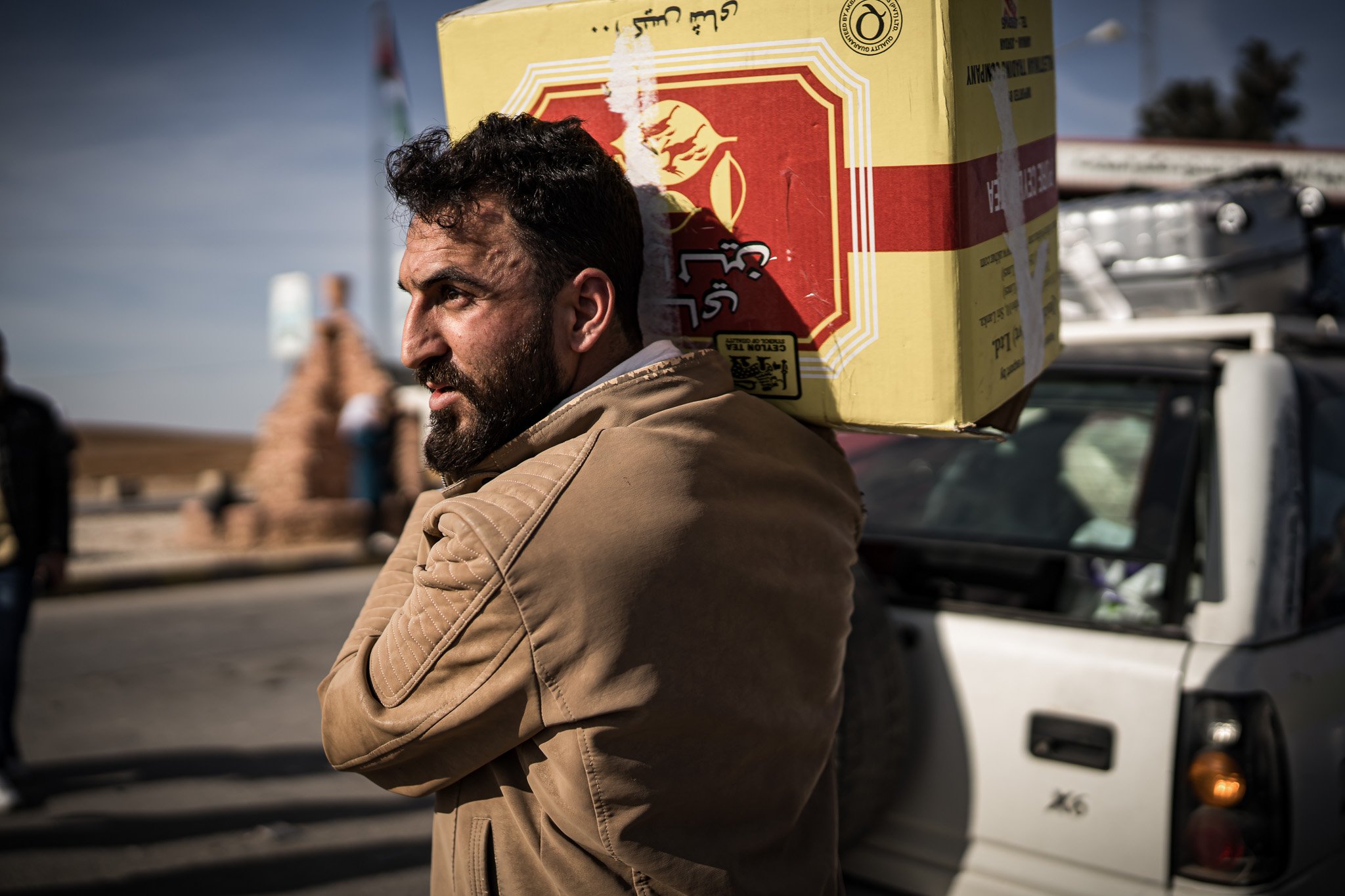 A man with a beard and tan jacket carrying a large box on his shoulder, standing in front of a vehicle and outdoor scene with a structure and people in the background.