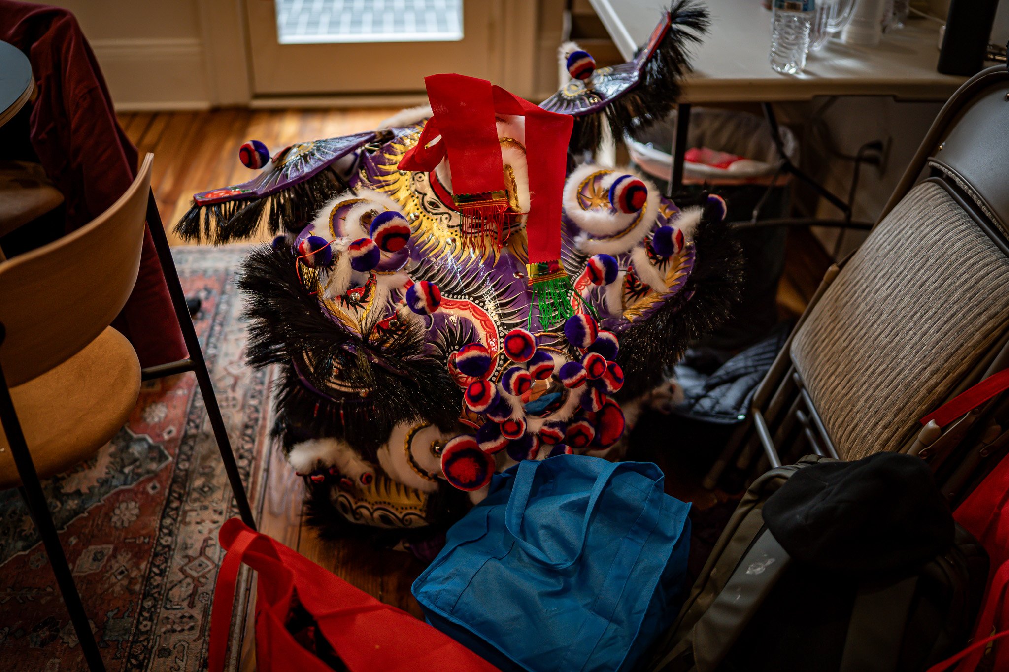 A colorful, decorated Chinese dragon costume with feathers and pom-poms, placed on a chair in a room with chairs, tables, water bottles, and a bag.