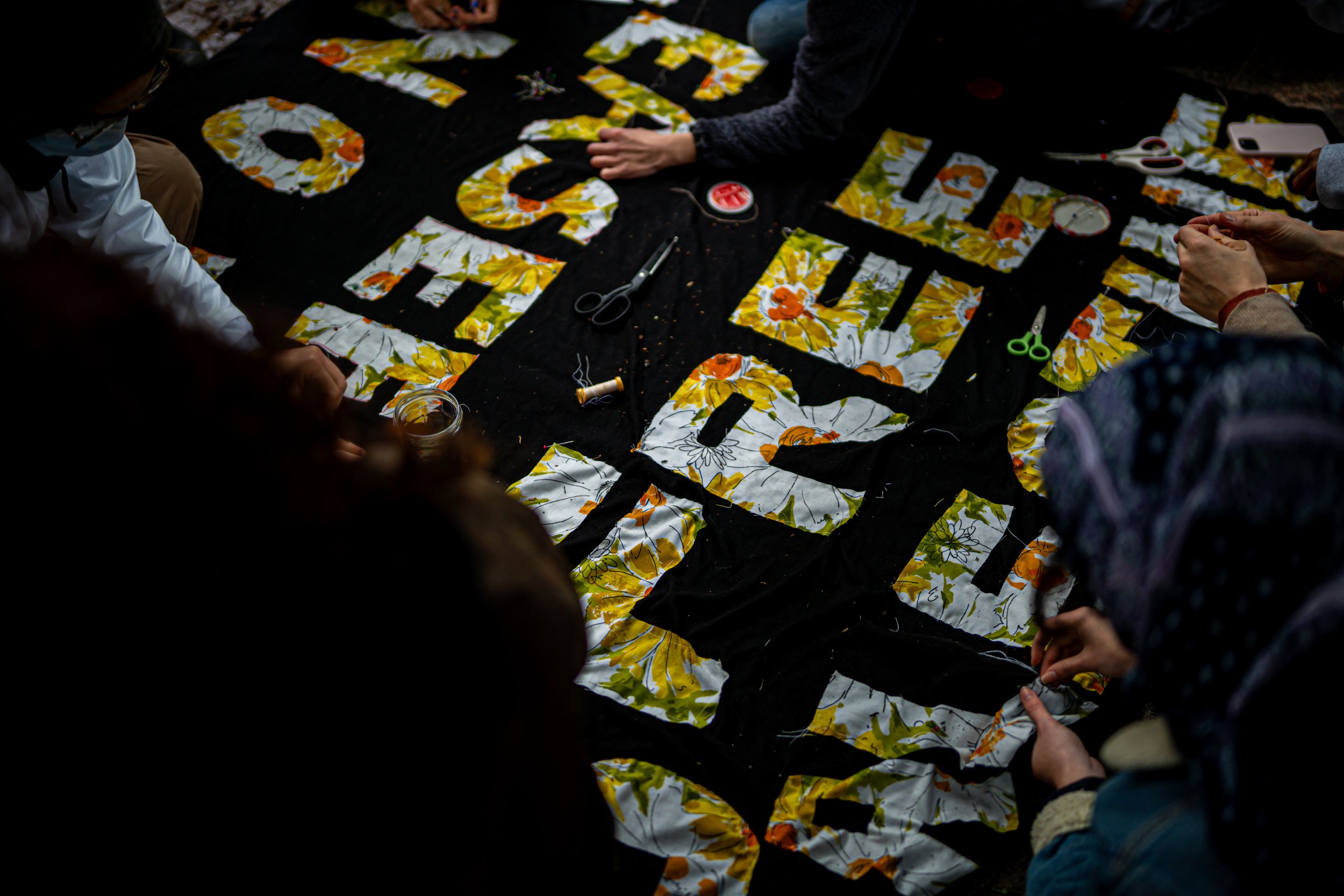 People gathered around a black cloth with large cut-out letters, working on a fabric banner with floral patterns and text.