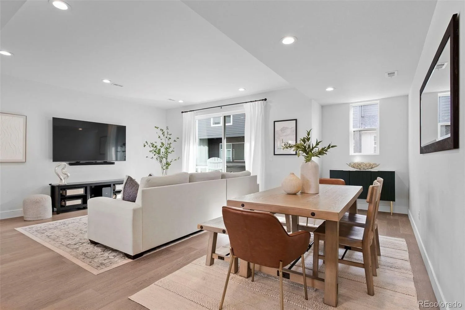 Modern living room with a white sofa, flat-screen TV, wooden dining table with chairs, and large windows with white curtains, decorated with plants and artwork.