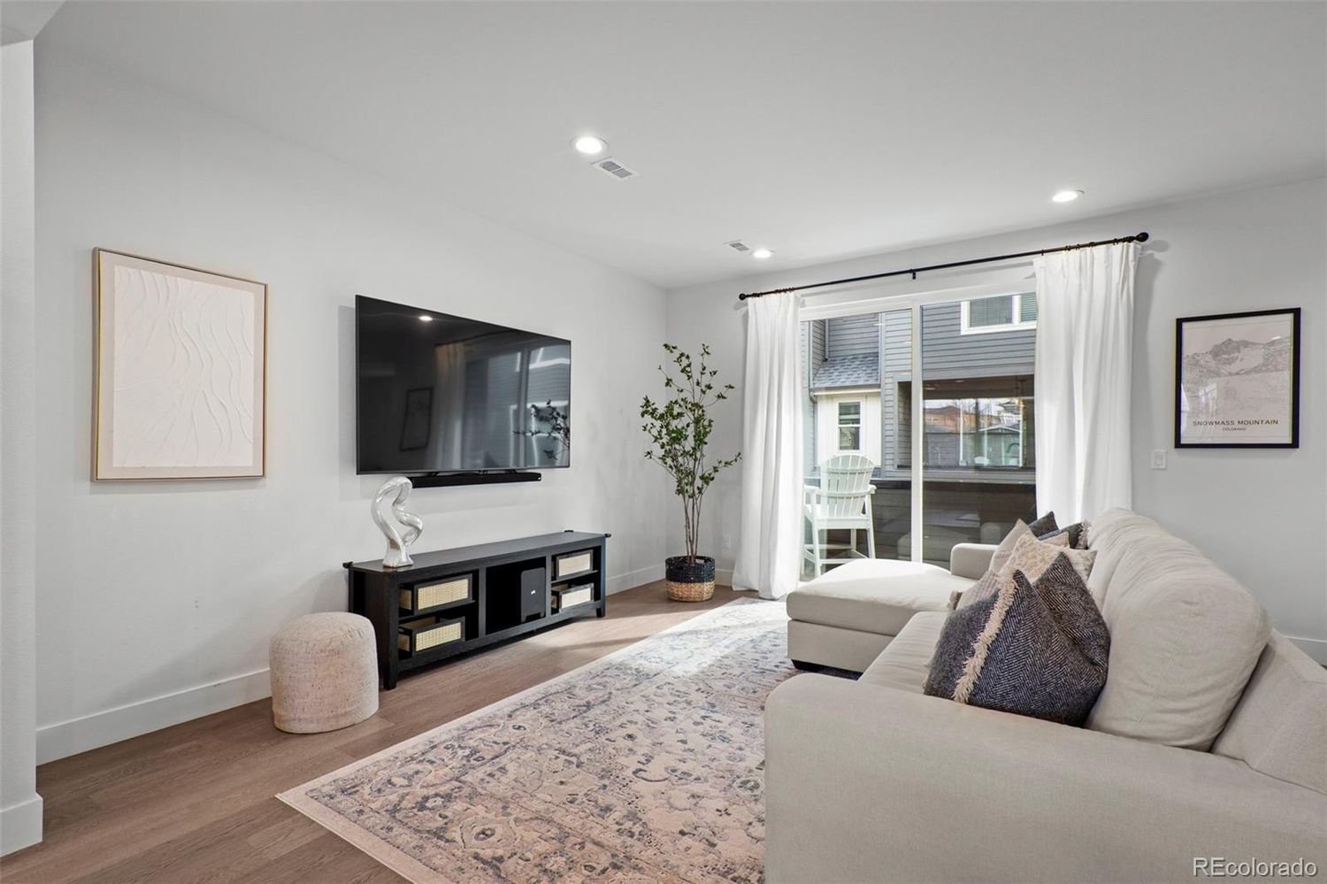 Bright living room with white walls, a beige sectional sofa with throw pillows, a patterned area rug, a flat-screen TV mounted on the wall, a black TV stand, a potted plant, and a sliding glass door with white curtains leading to a balcony with outdo