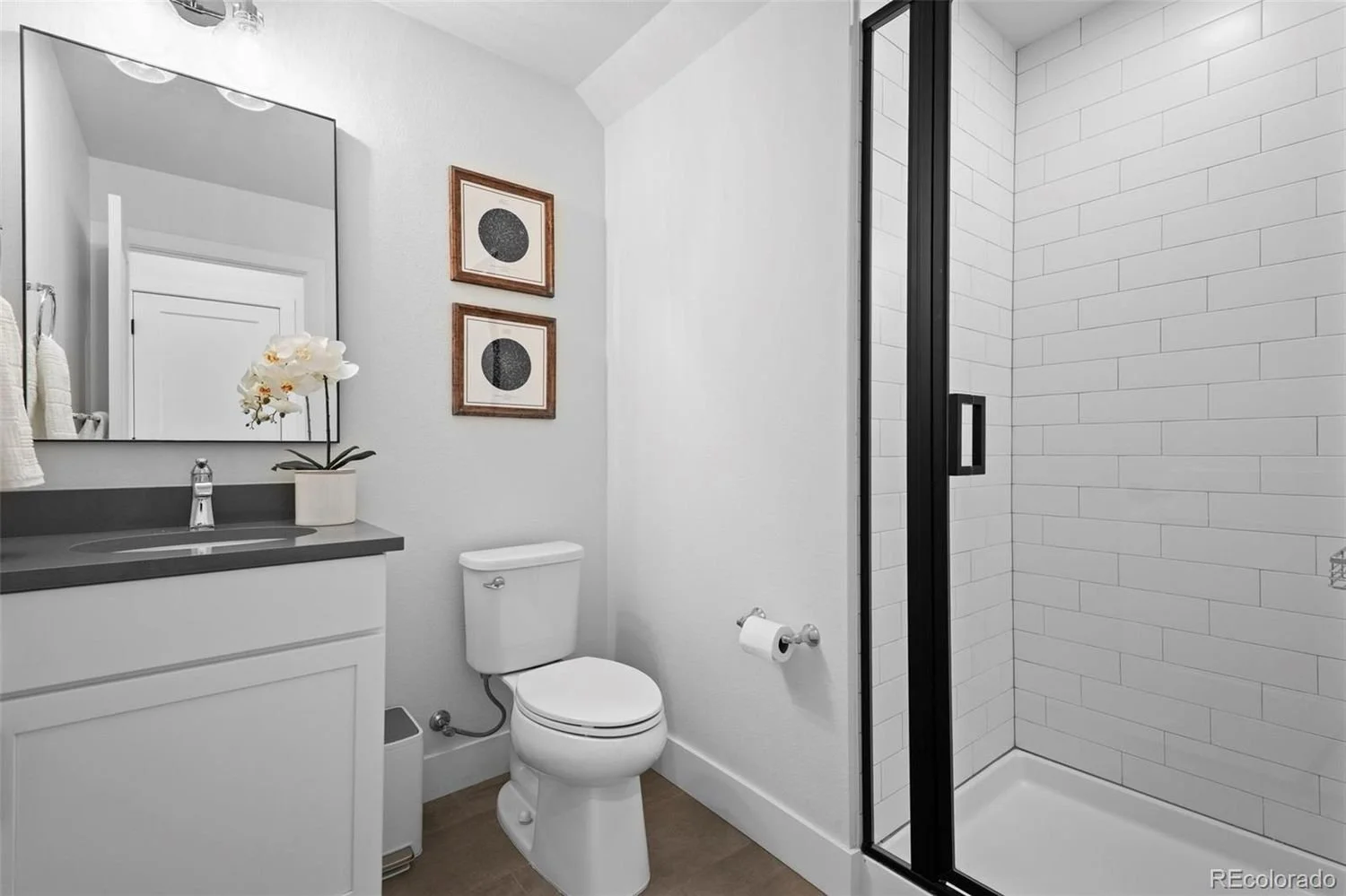 Modern bathroom with white walls, a toilet, a vanity with a dark countertop, a mirror, decorative framed art, and a glass-enclosed shower with white subway tiles.