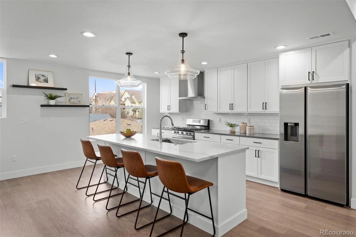 Modern white kitchen with stainless steel refrigerator, oven, white cabinets, pendant lights, and a large island with four brown chairs.