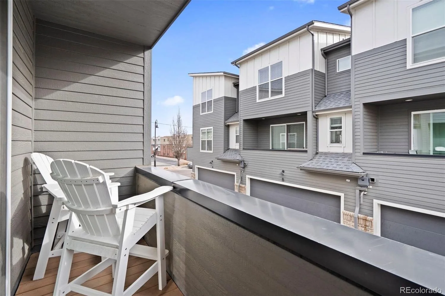 Small balcony with two white Adirondack chairs overlooking neighboring gray and white townhouses with garage doors, windows, and small balconies, in a suburban area on a clear day.