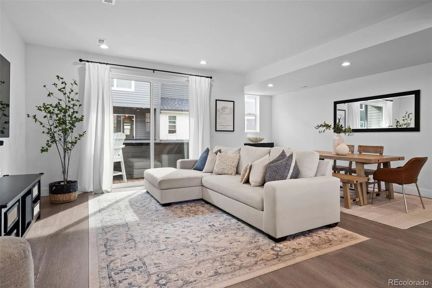 Living room with white L-shaped sofa, throw pillows, potted plant, large window with white curtains, framed art, wooden dining table with chairs, and a large rectangular mirror on the wall.
