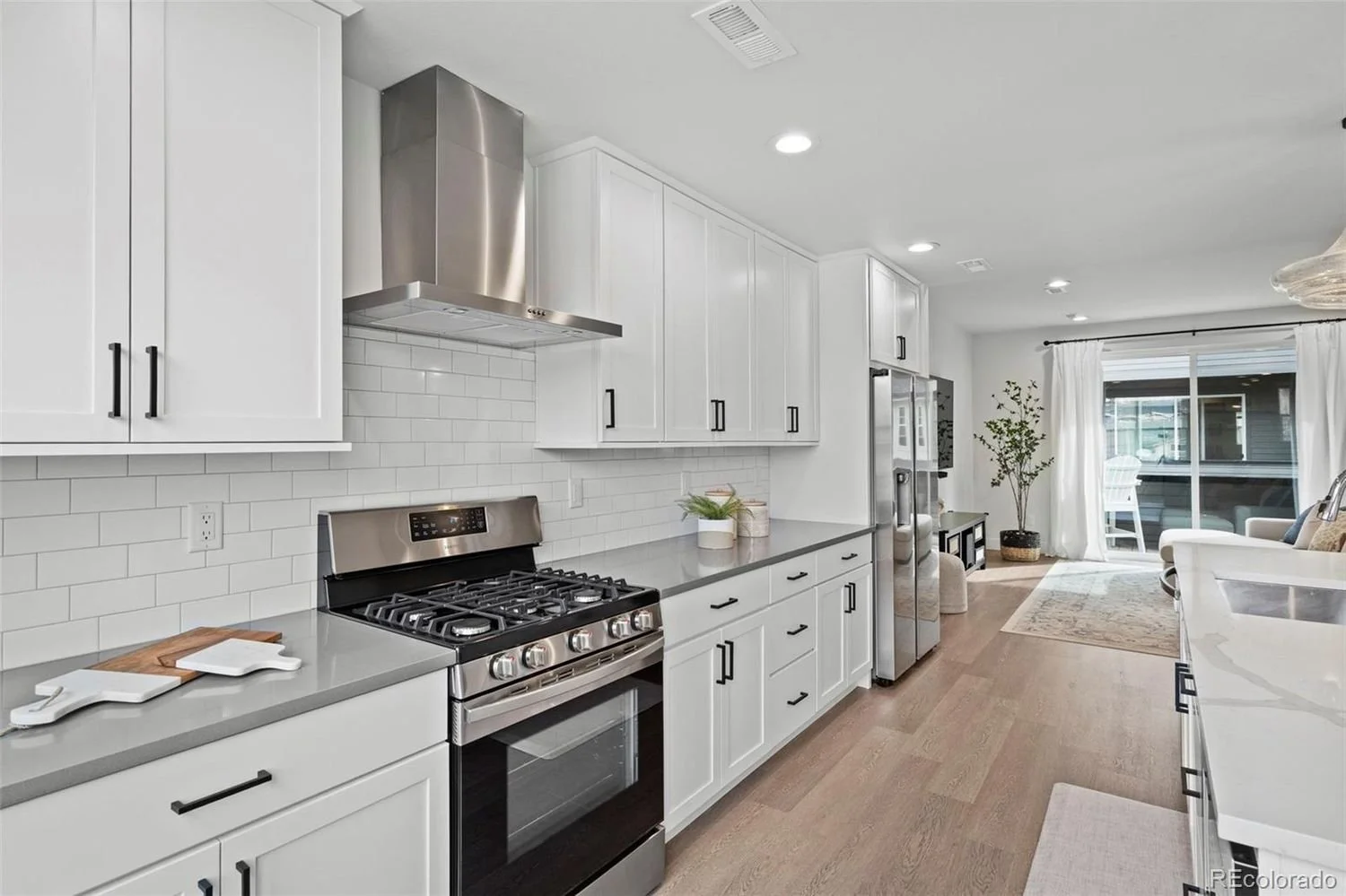 Modern kitchen with white cabinets, gray countertops, stainless steel appliances, white subway tile backsplash, potted plant, and a view into a living area with sliding glass door and white curtains.