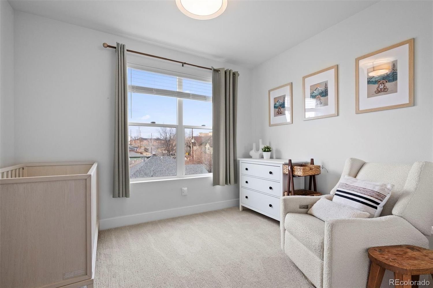 A simple, bright nursery room with a large window, beige carpeting, a white dresser, an armchair with a pillow, and wall art featuring animals and mountain scenery.