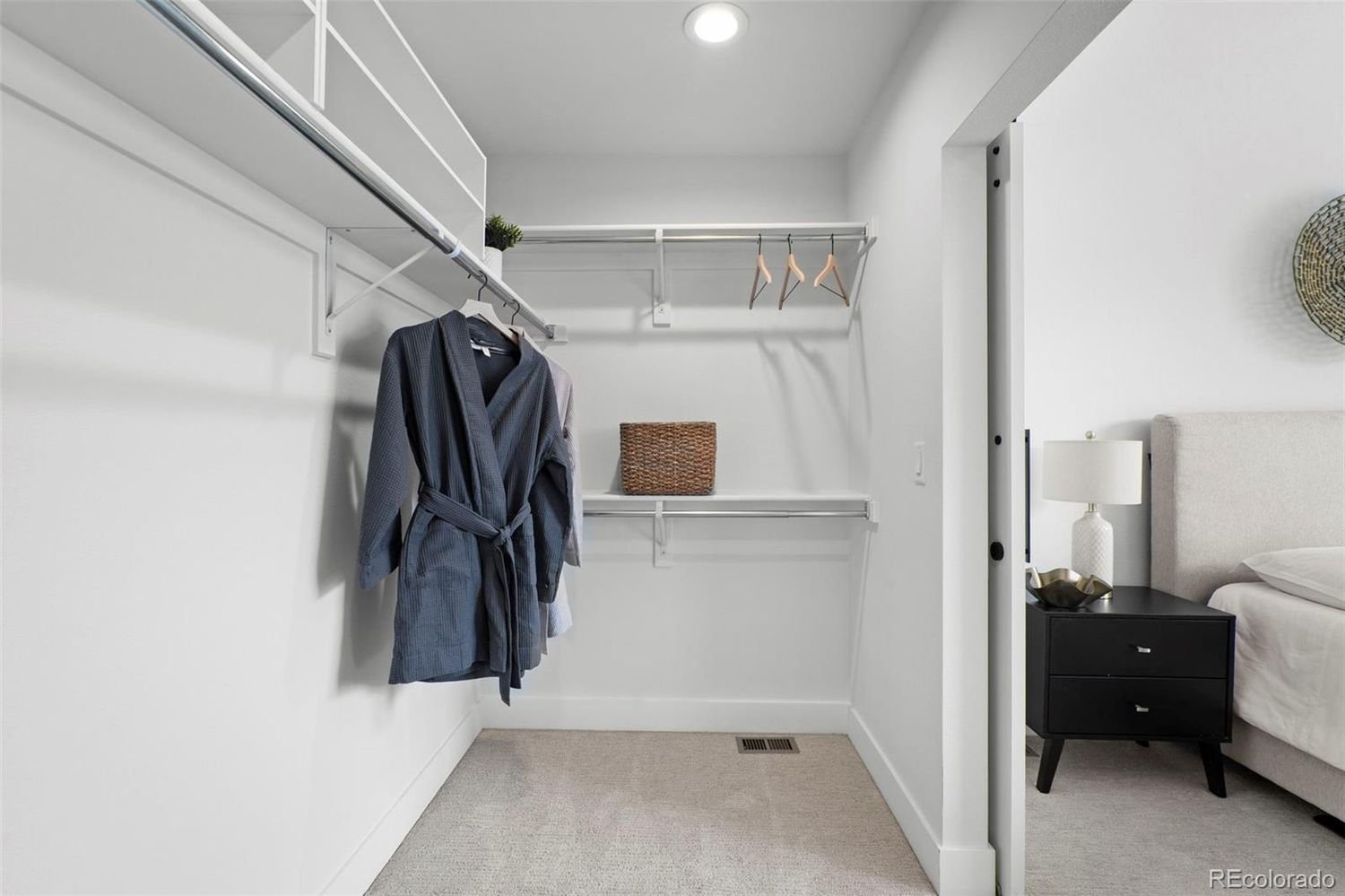 Empty walk-in closet with white walls, a beige carpet, and a few clothes on a hanging rod, with a bedroom visible to the right.