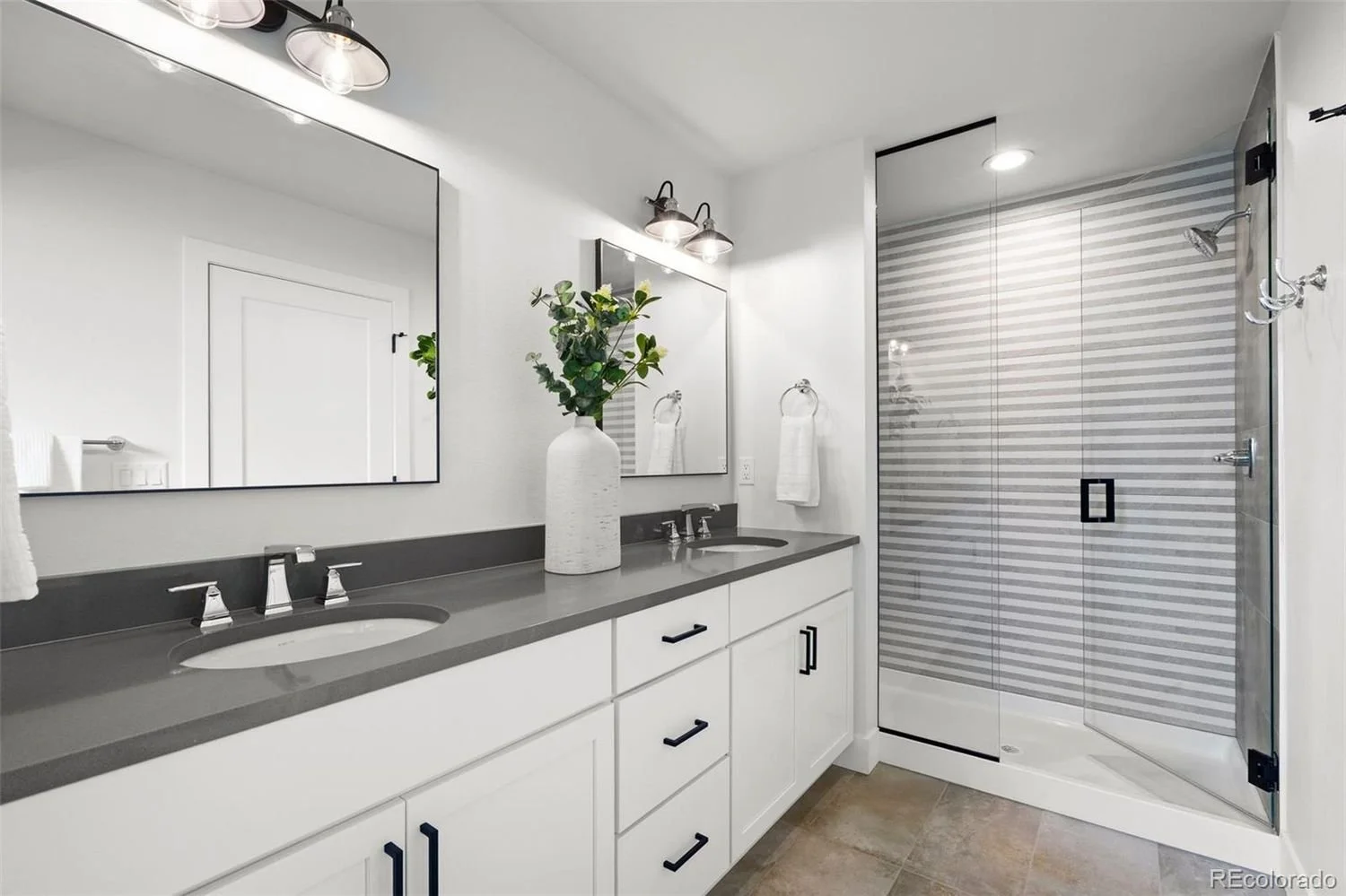 Modern bathroom with double vanity, gray countertop, white cabinets, black handles, large framed mirrors, a vase with greenery, wall-mounted light fixtures, a glass-enclosed shower with striped pattern glass, and beige tile flooring.