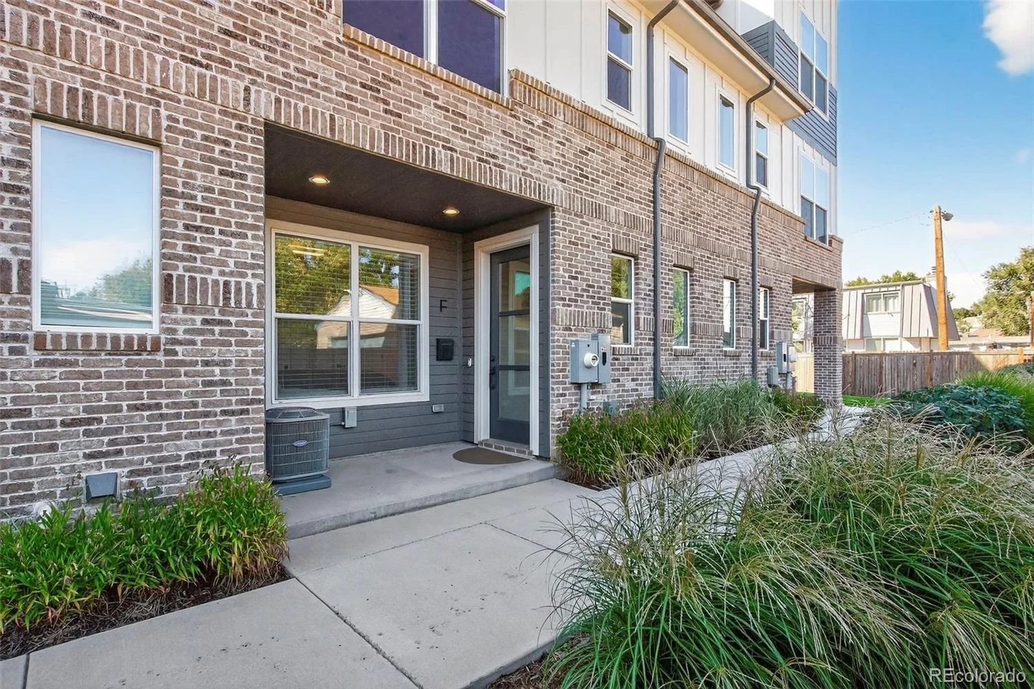 Exterior view of a modern apartment building with a brick facade, large windows, and small front porch with plants and sidewalk.