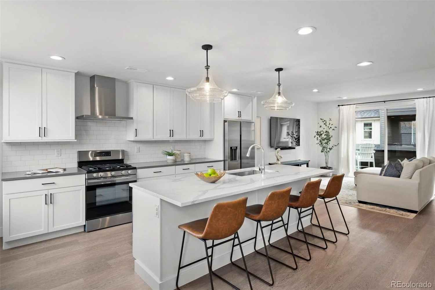 Modern white kitchen with an island, four brown leather chairs, and stainless steel appliances, open to a living room with a white sofa and sliding glass door.