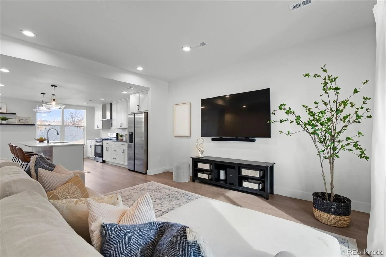 Living room with a beige sofa, decorative pillows, a large flat-screen TV on the wall, a black TV stand, a potted plant, and a view of the kitchen in the background with white cabinets and a dining area with barstools.