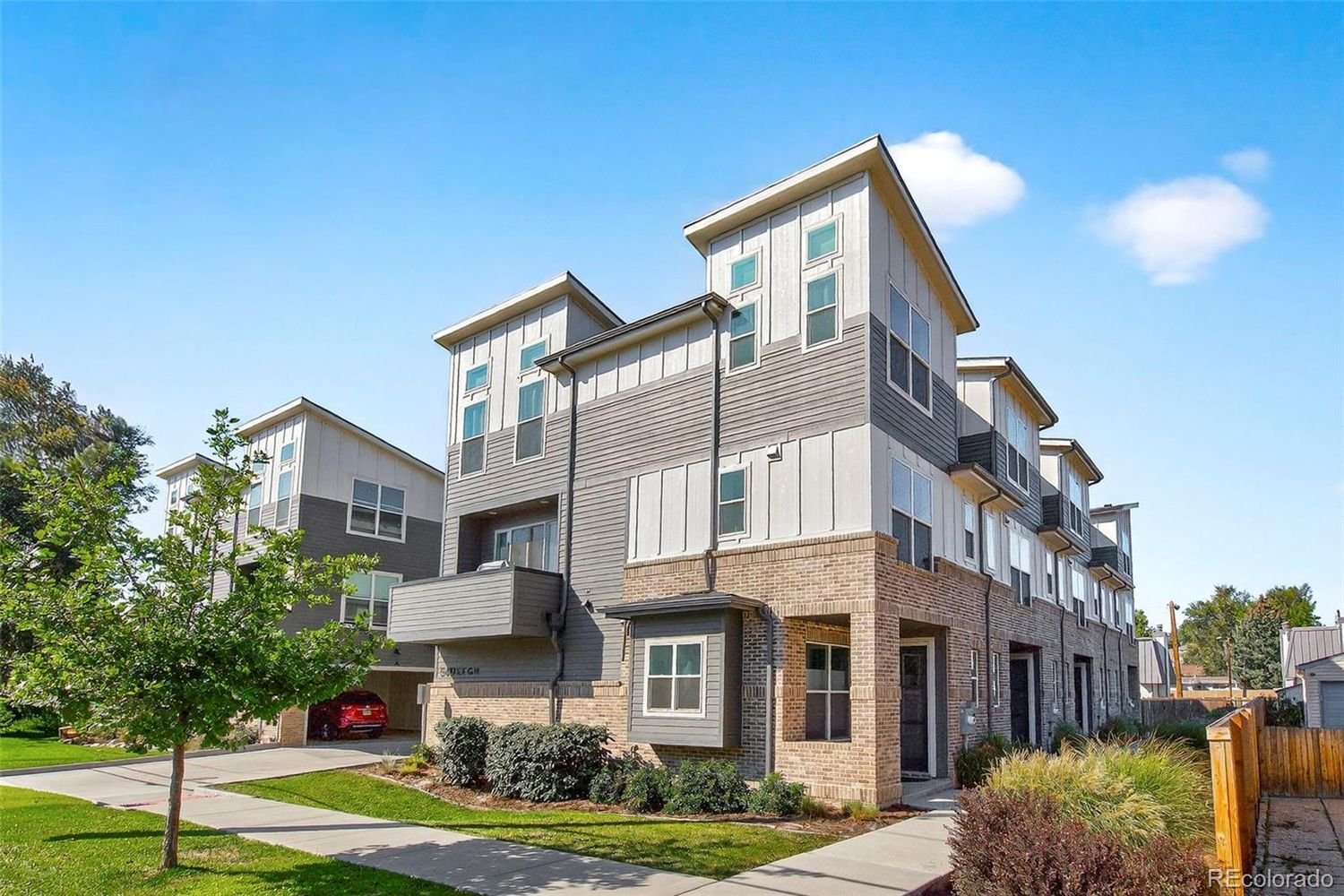 Multi-story residential building with gray and white exterior, brick foundation, and multiple windows, surrounded by a green lawn, bushes, and a small tree under a clear blue sky.