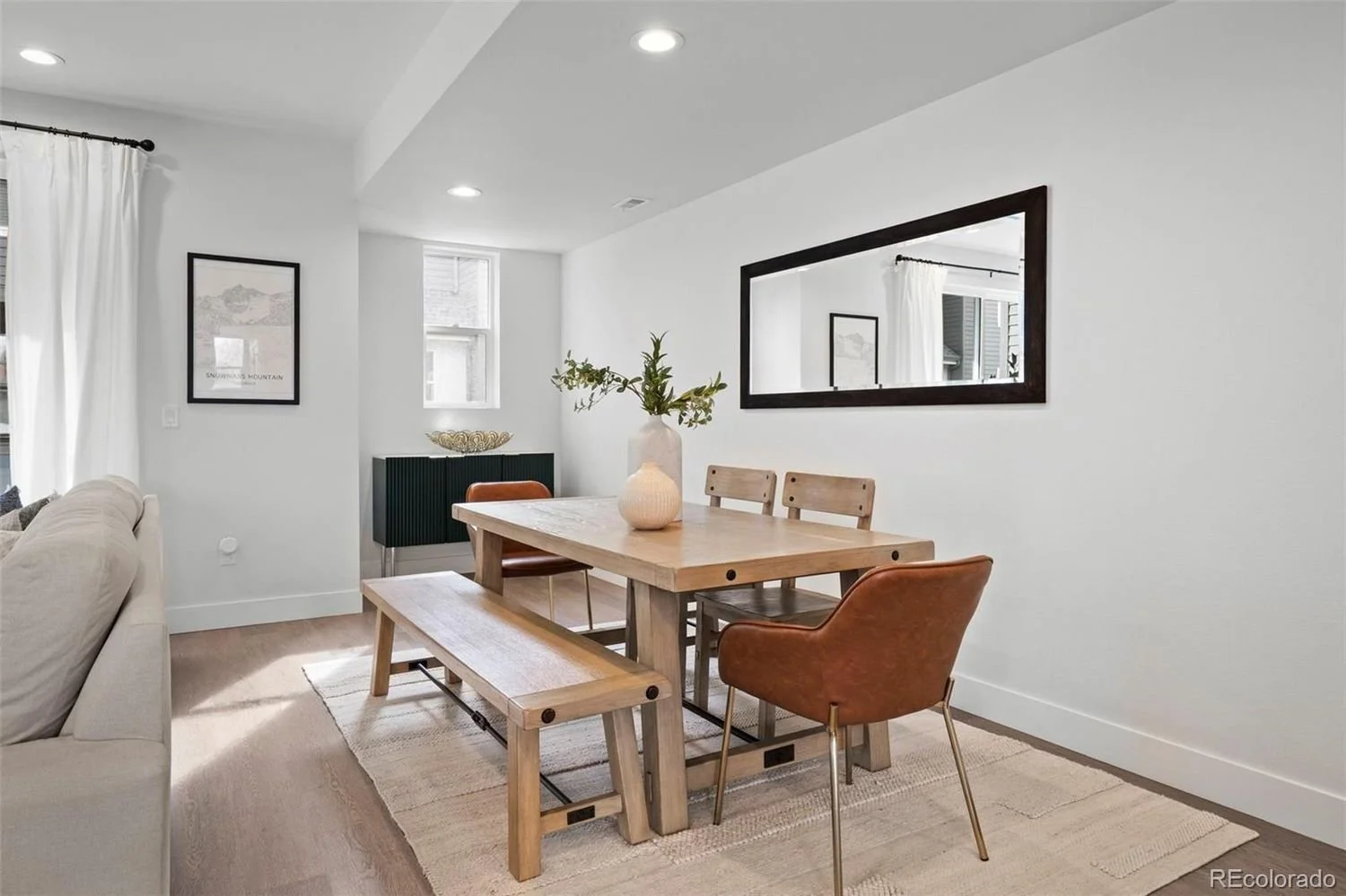 Dining room with wooden table, brown chairs, beige bench, large mirror on white wall, decorative vases and picture on wall, windows with white curtains, beige carpet, and light-colored walls.
