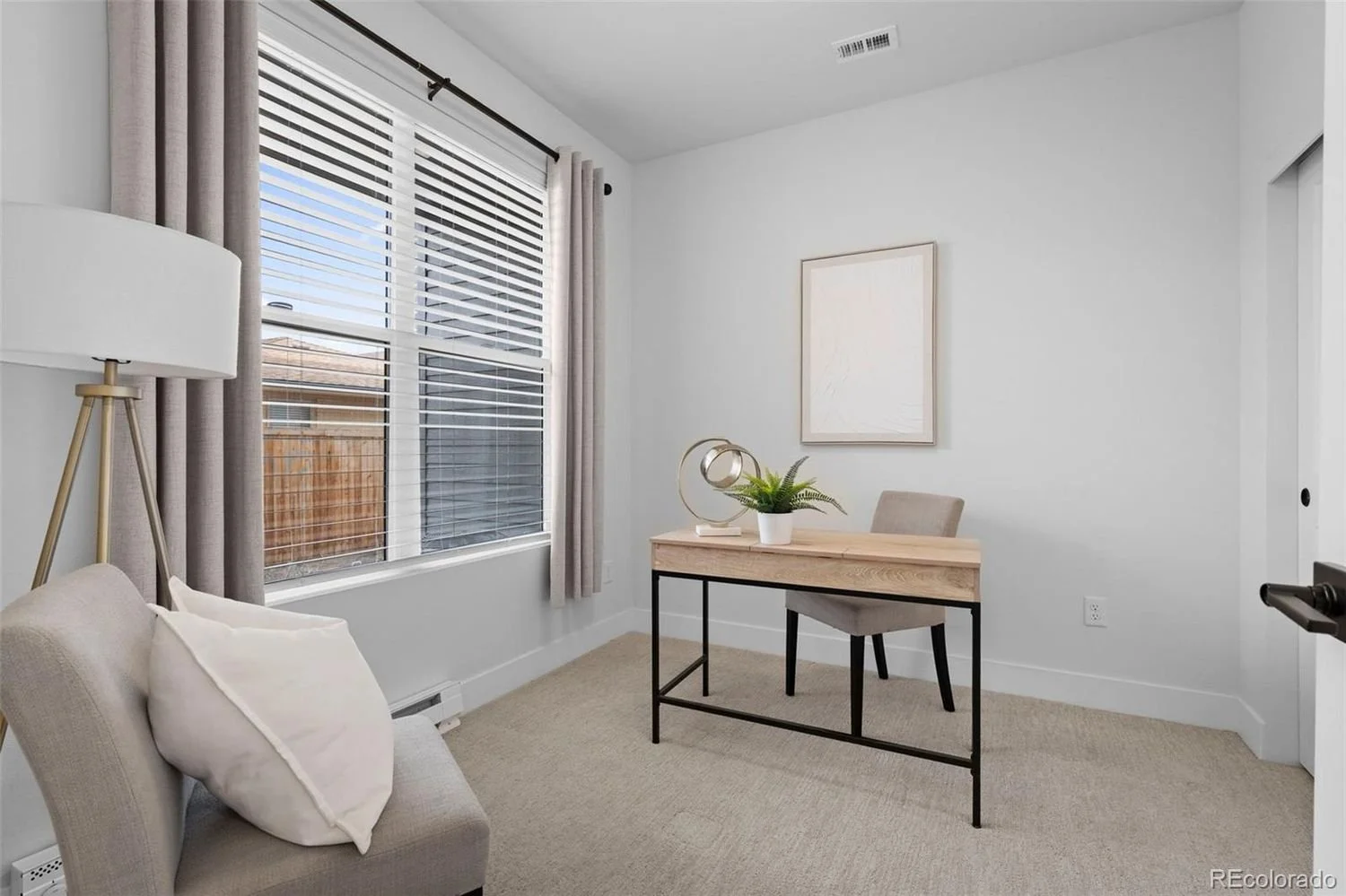 A small, minimalistic home office with a window, curtains, a desk with a potted plant and lamp, a chair, and a abstract artwork on the white wall.