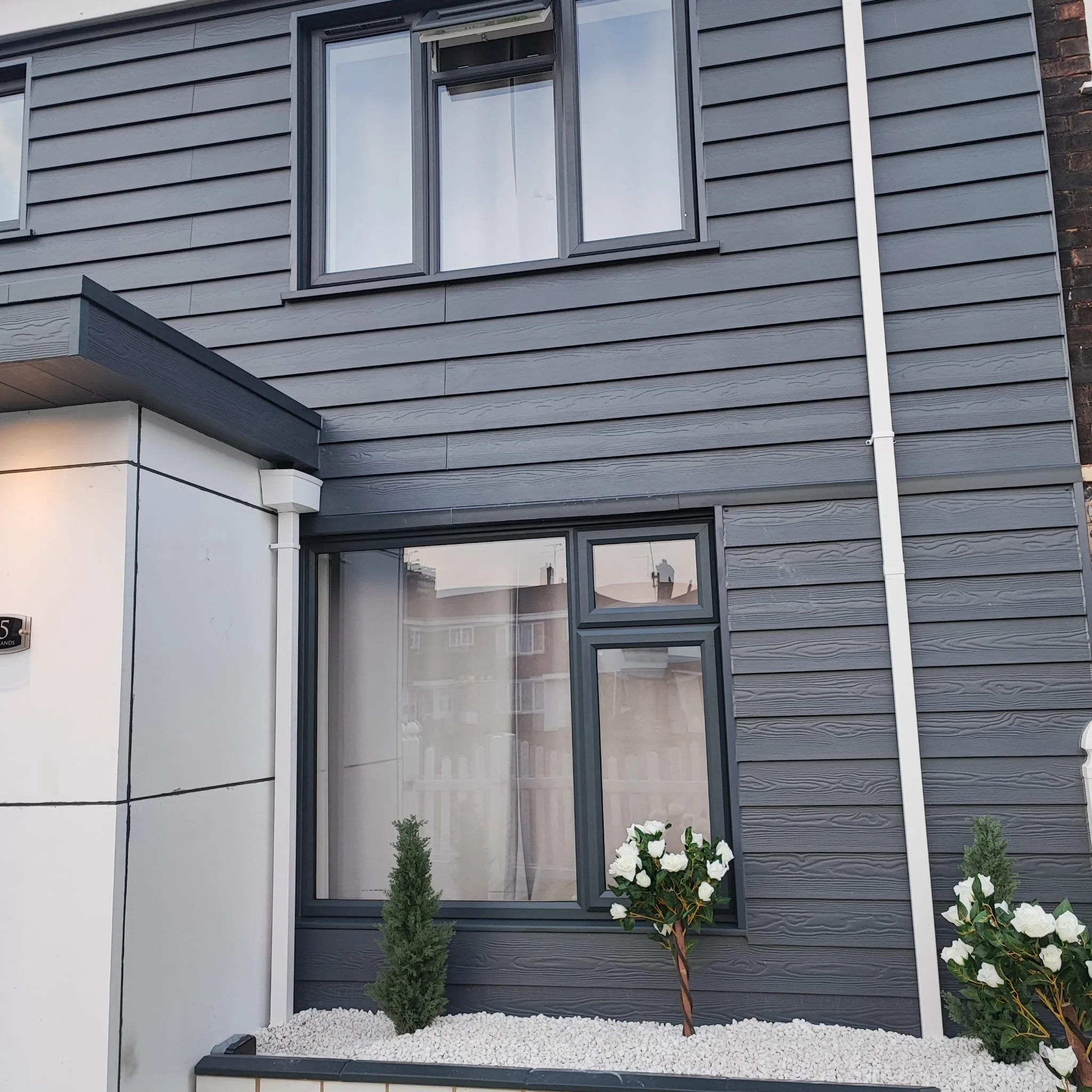Close-up of a modern house's exterior showing dark gray horizontal siding, a large window, and a small garden with white gravel, green shrub, and flowering plant.