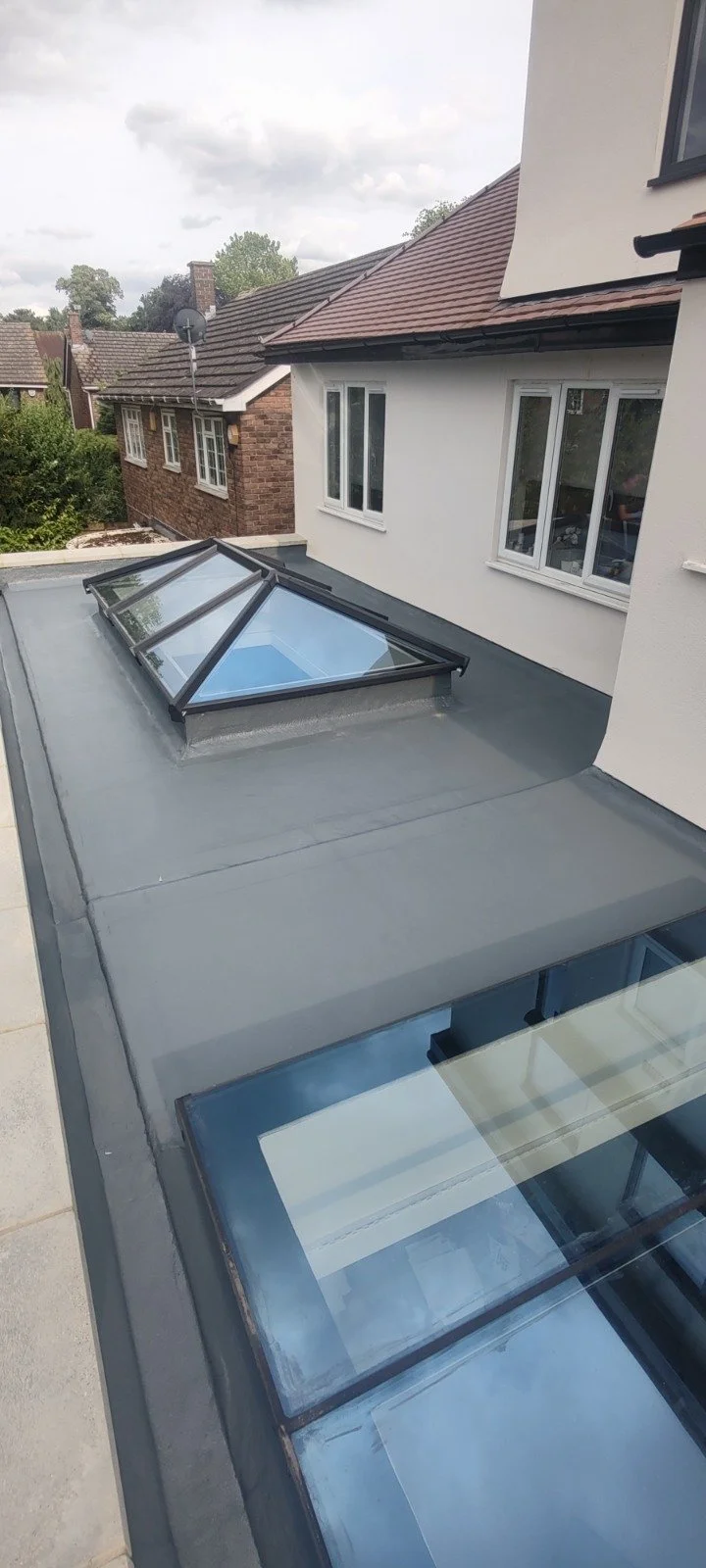 View of a rooftop with a skylight enclosed in a glass pyramid structure on a gray roof, part of a white house with multiple windows.