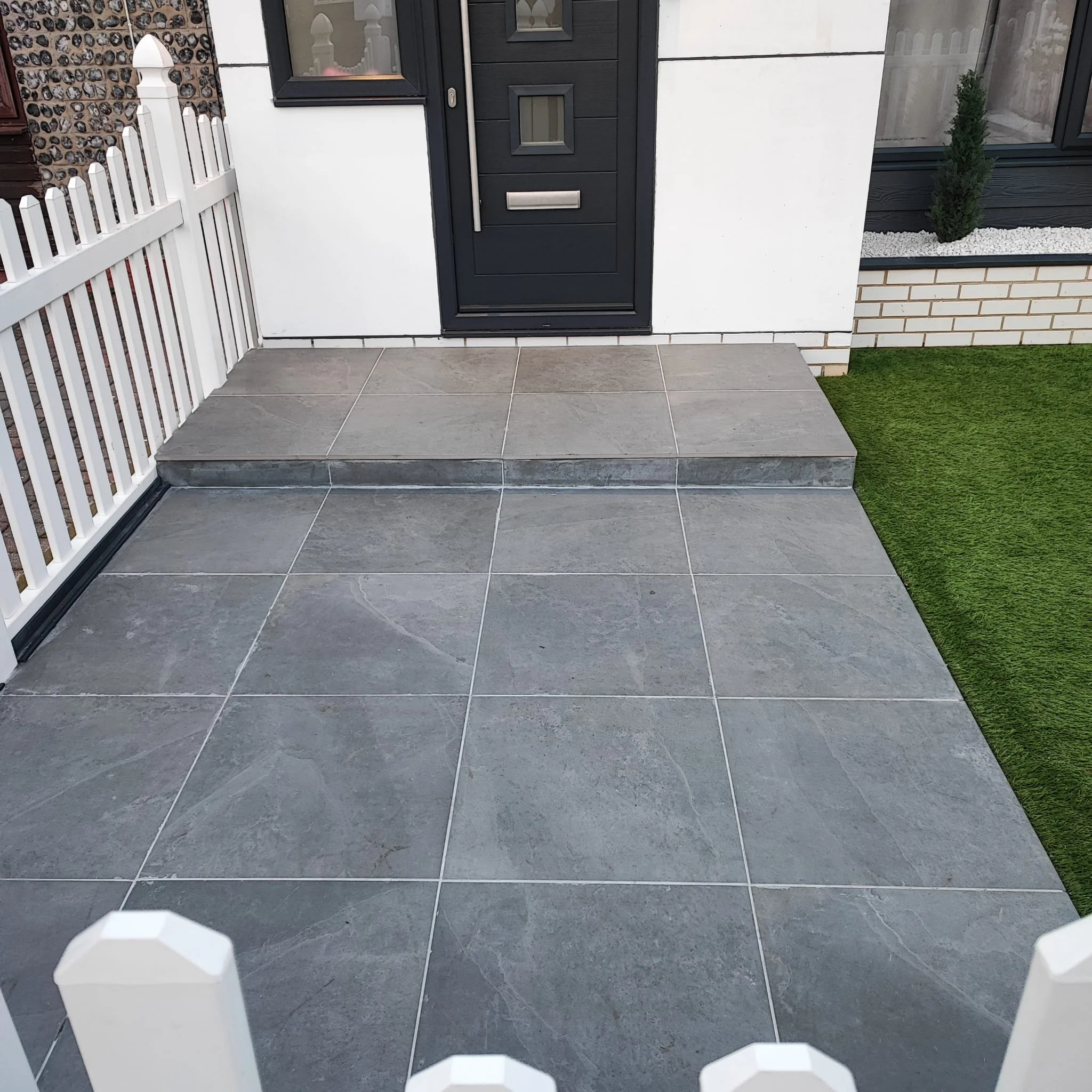 Front entrance of a modern house with gray tiled porch, black front door with small windows, white fence on the left, and a section of artificial grass on the right.