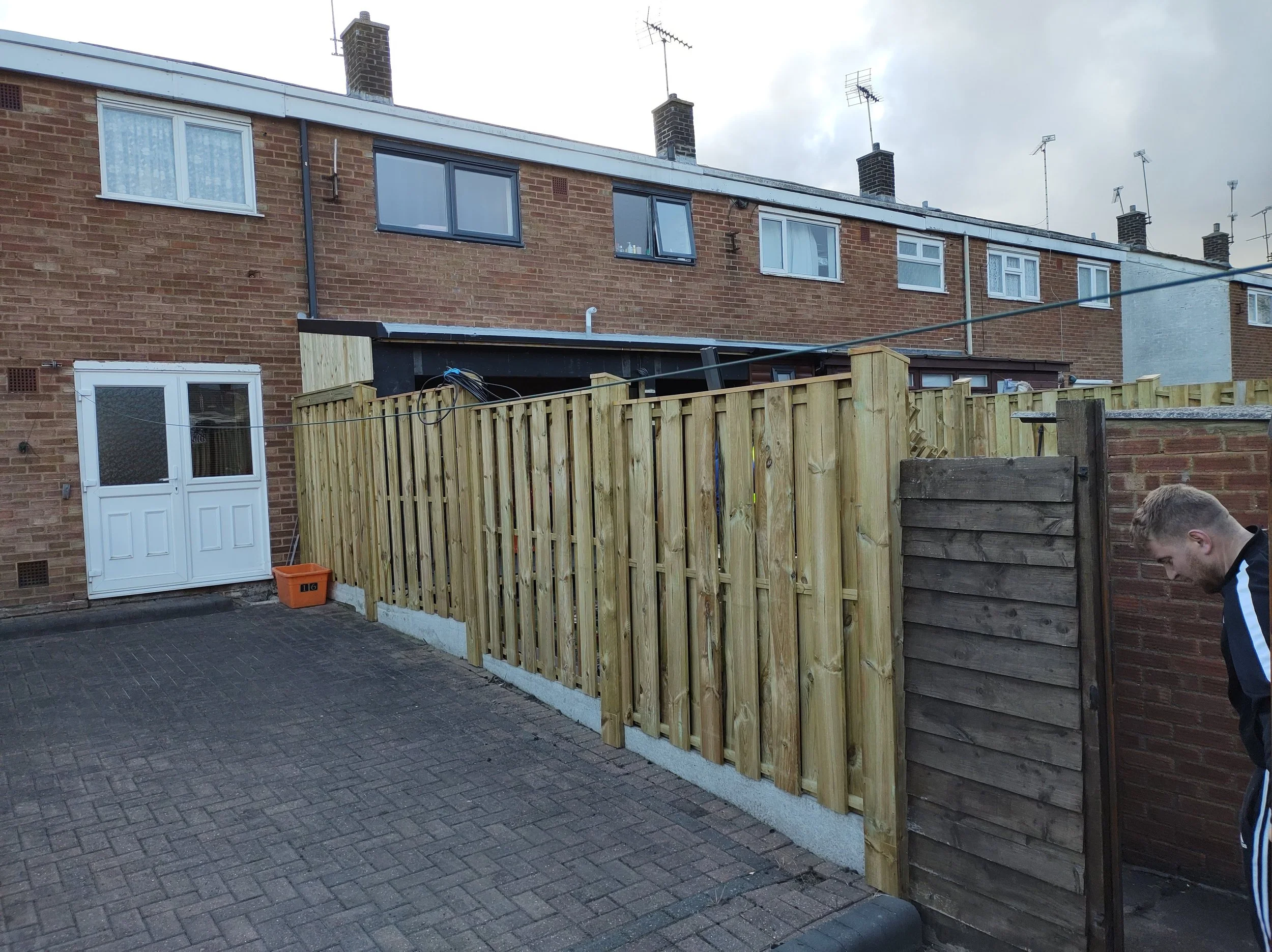 A backyard with a new wooden privacy fence, brick houses in the background, and a person in a black and white jacket leaning on the fence to the right.