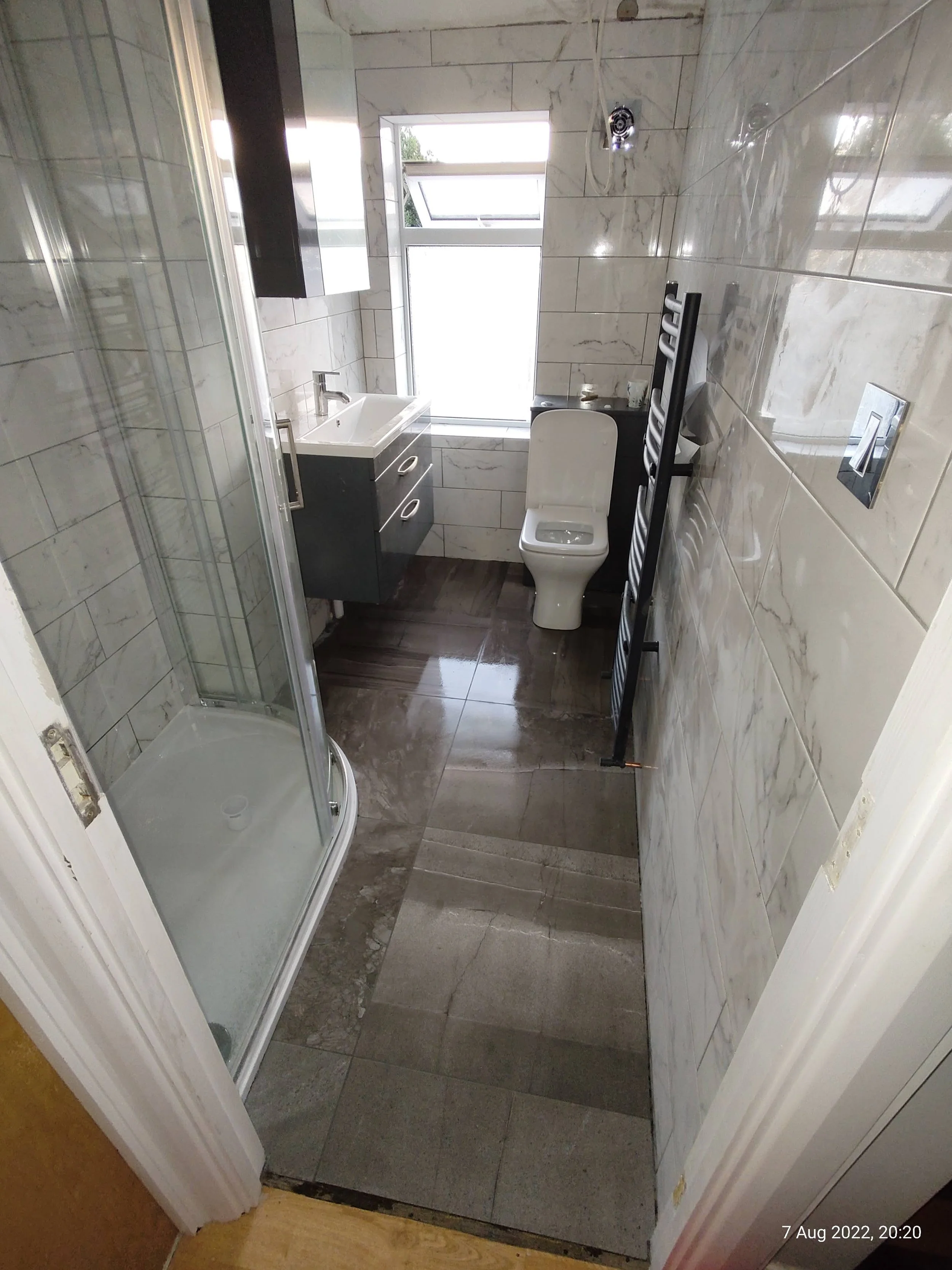 Small modern bathroom with a glass shower enclosure, a white toilet, a dark vanity with a sink, a window, and a heated towel rack on a tiled wall.