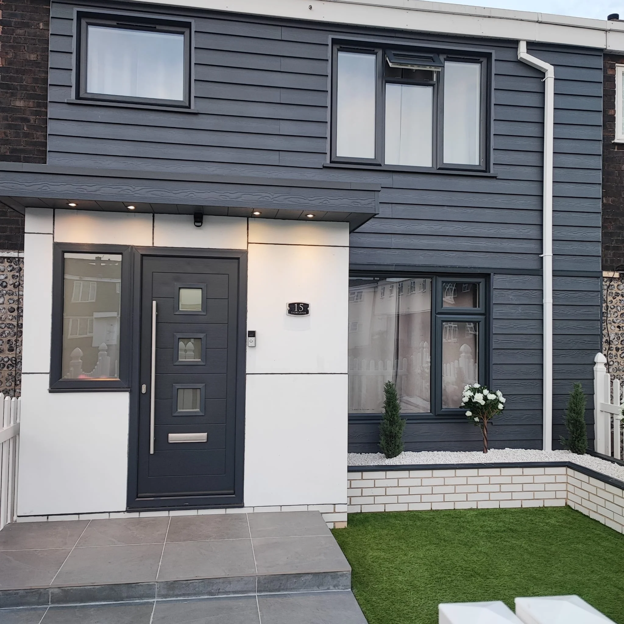 Modern two-story house with black siding, white accents, and a black front door with square windows, small plants, and a white picket fence.