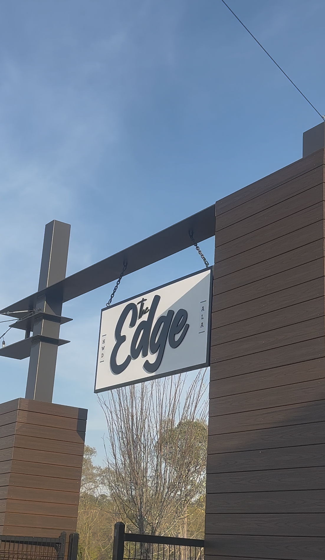 A sign that reads 'The Edge' hanging from a black metal frame against a blue sky, with wooden panel walls and some trees in the background.