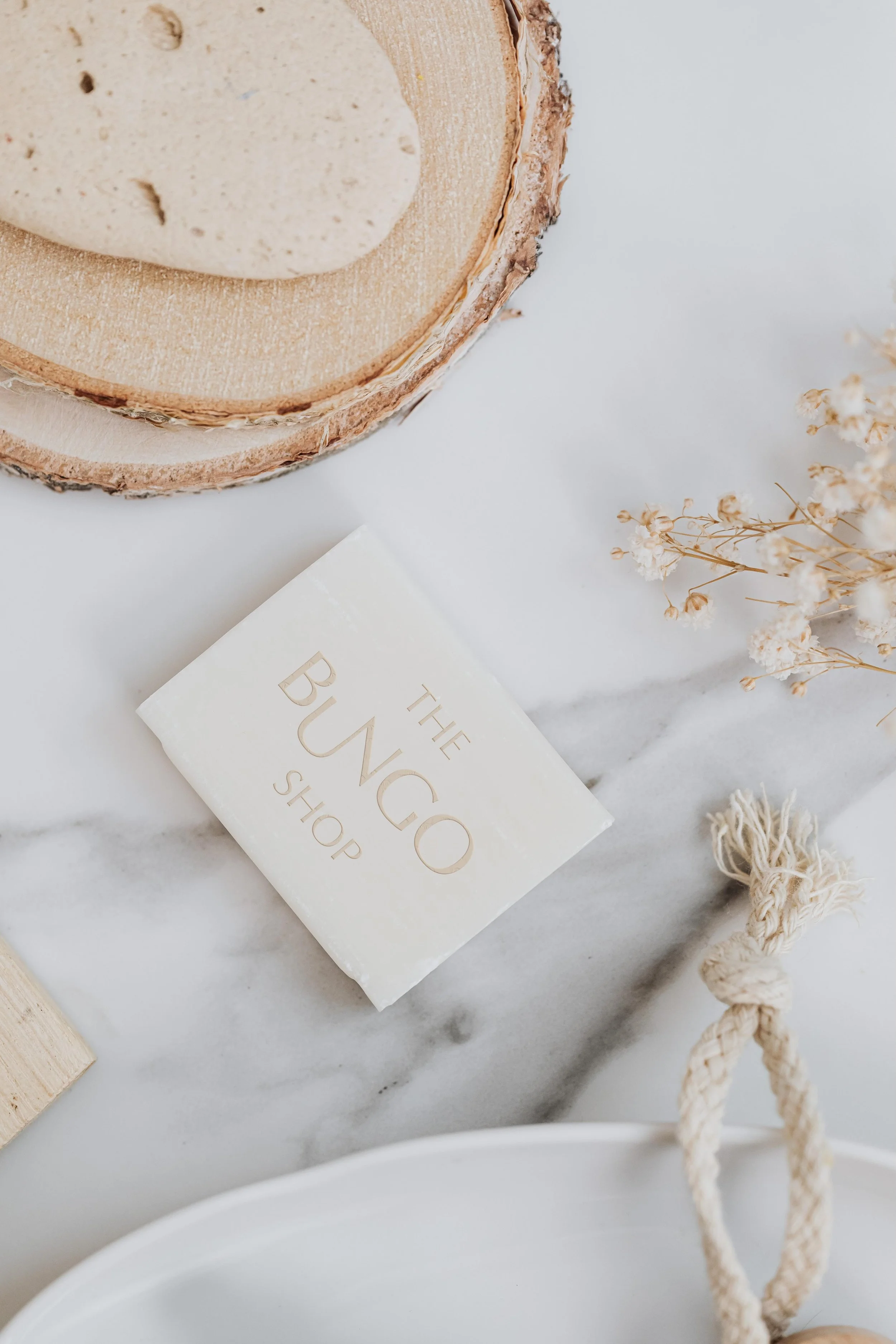 Close-up of a wooden soap dish with a bar of beige soap, a white soap box labeled 'THE BUCKE SHOP', small white flowers, and a beige braided rope on a white marble surface.