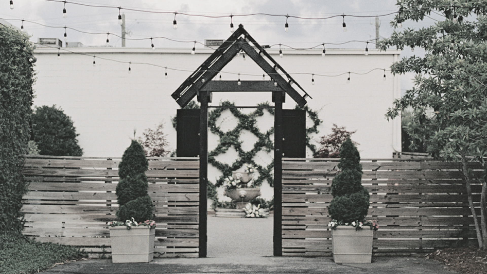 An outdoor garden scene with a black wooden arch adorned with greenery, surrounded by potted plants and a wooden fence, decorated with string lights overhead.
