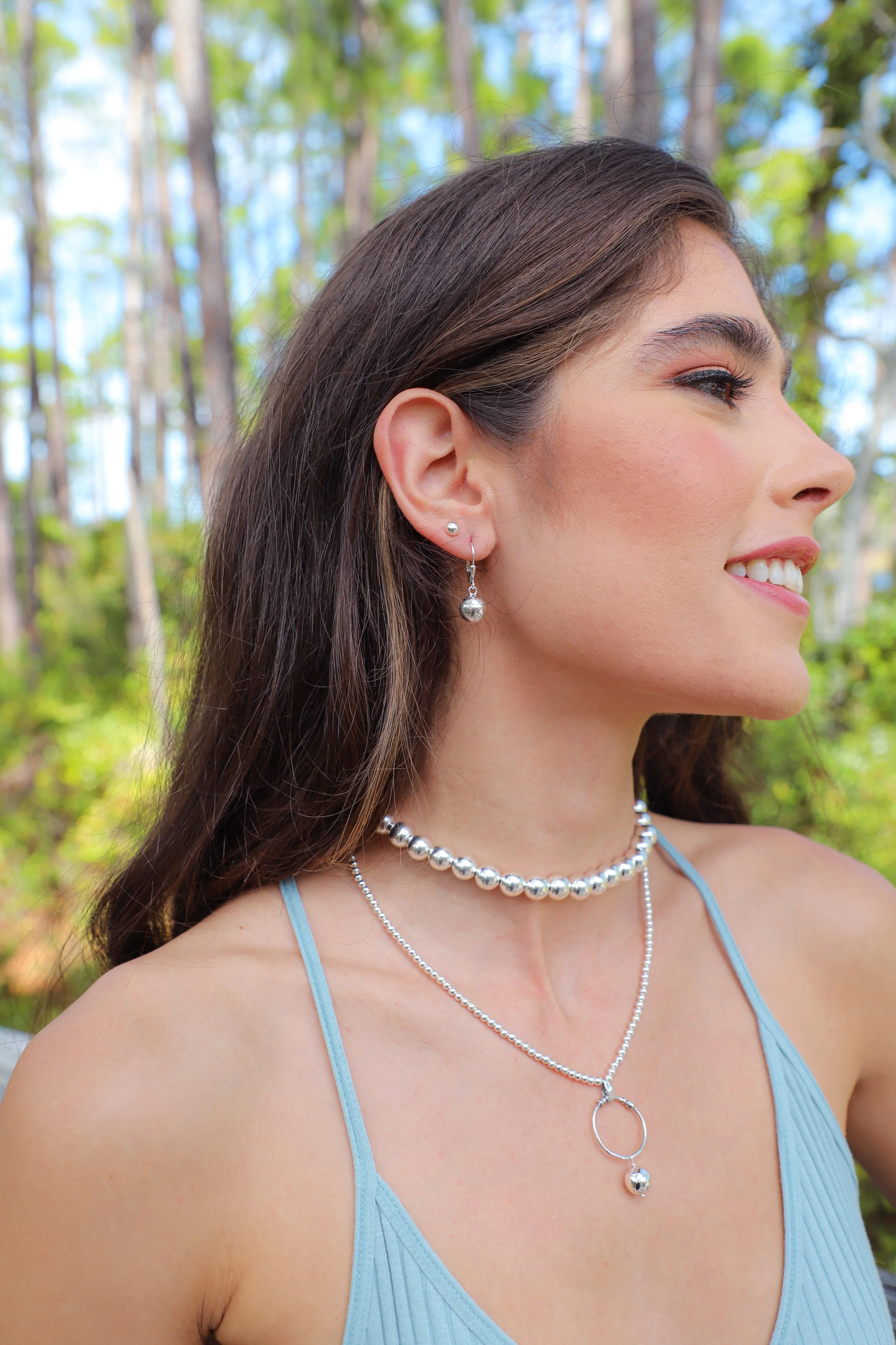 Profile of a young woman with long brown hair, wearing silver jewelry, and a blue dress, outdoors with a blurred background of green trees.