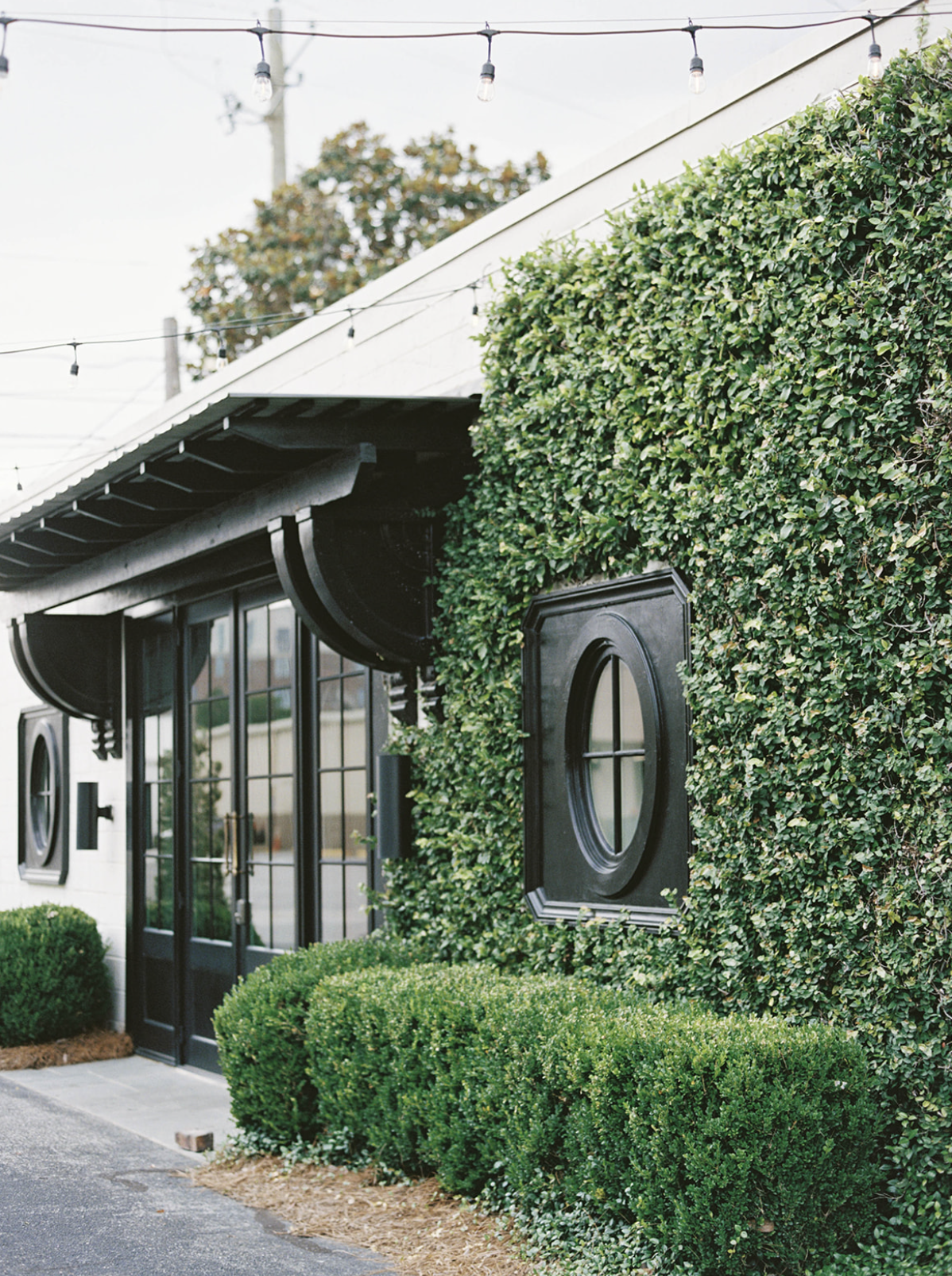 Exterior of a building with green ivy covering part of the wall, black decorative window frames, glass doors, and hanging outdoor string lights.