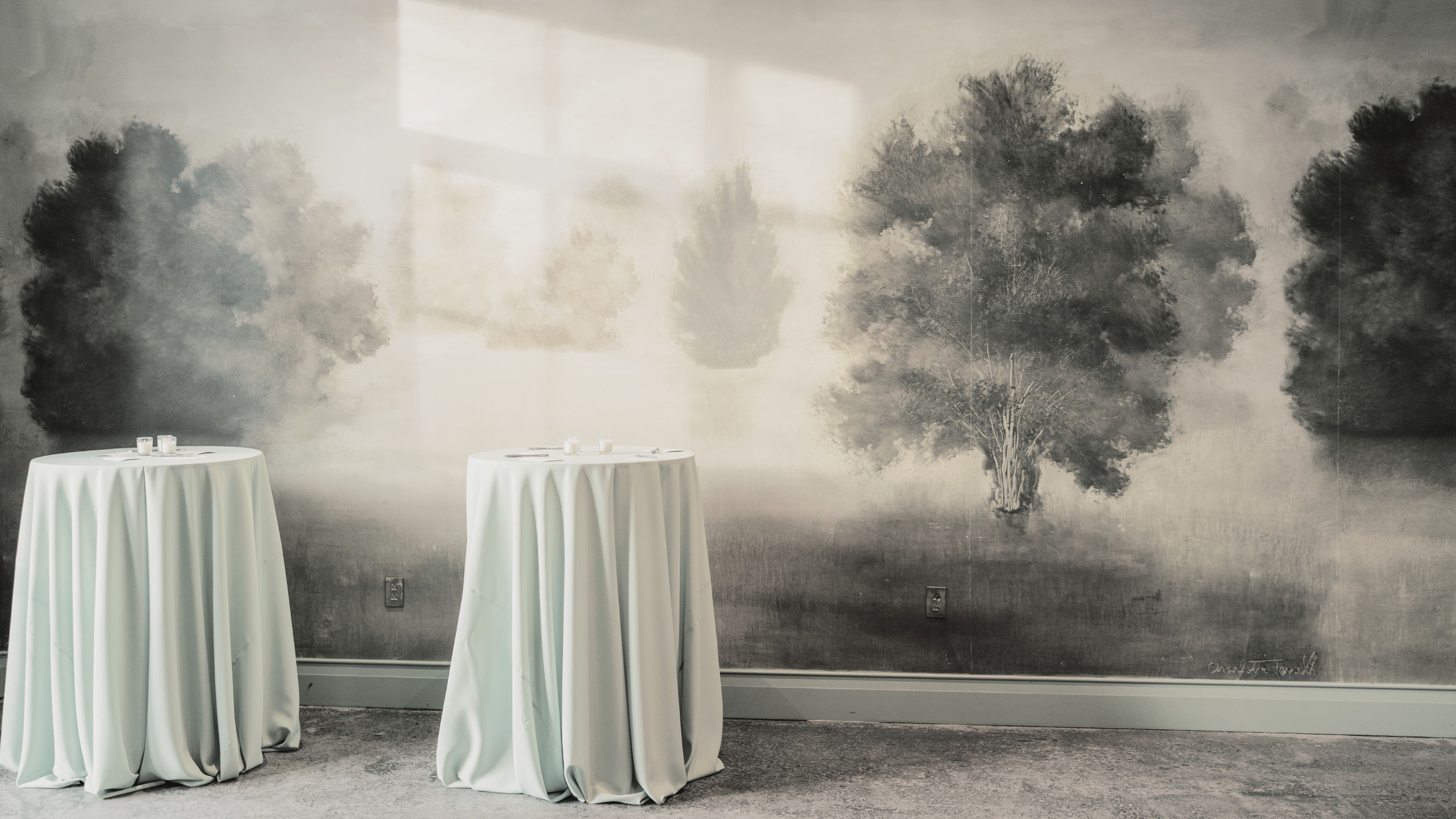 Two tall round tables with white tablecloths in front of a gray wall with a black-and-white watercolor painting of trees.